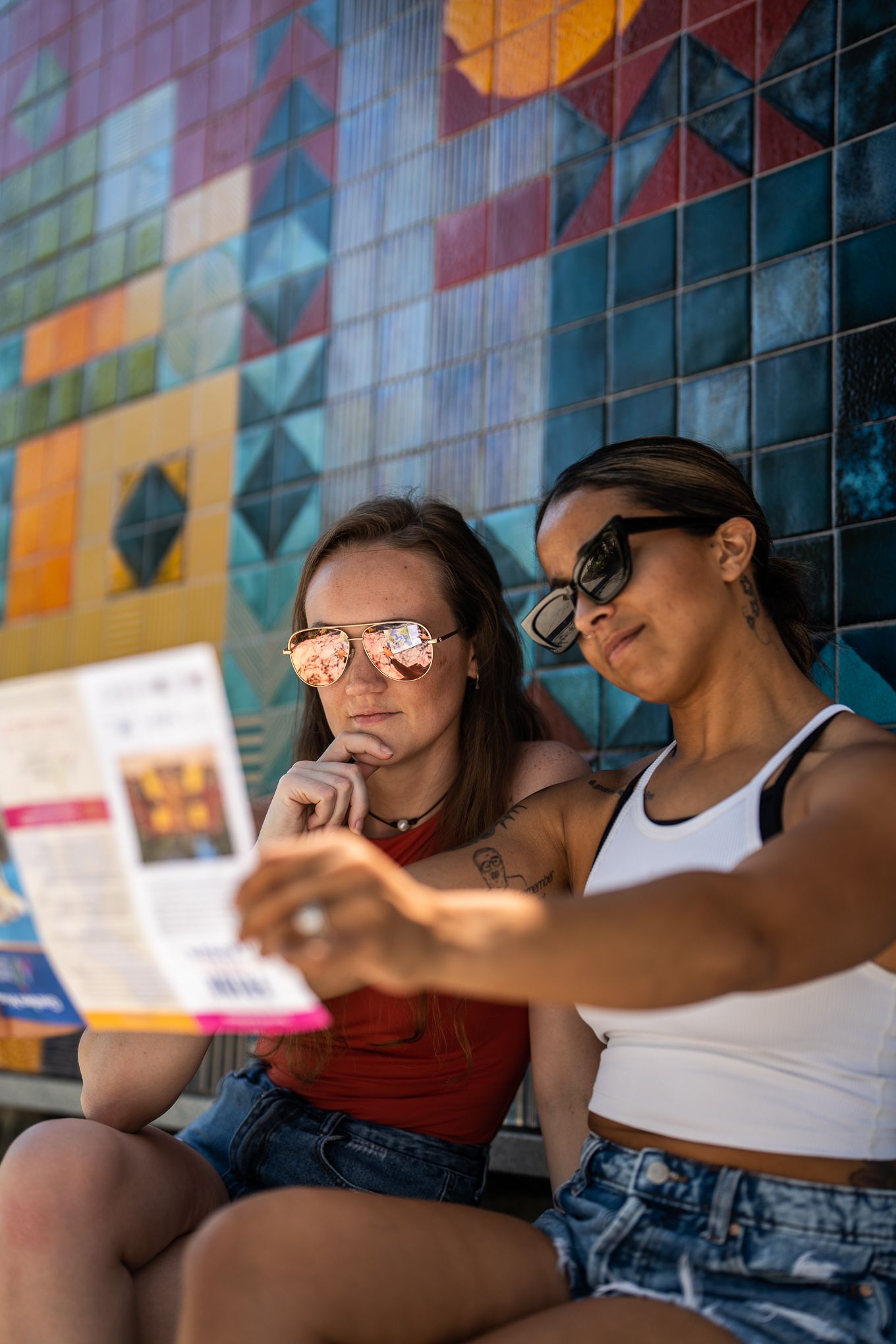 Two young women wearing sunglasses reading a travel brochure against a colorful tiled wall.