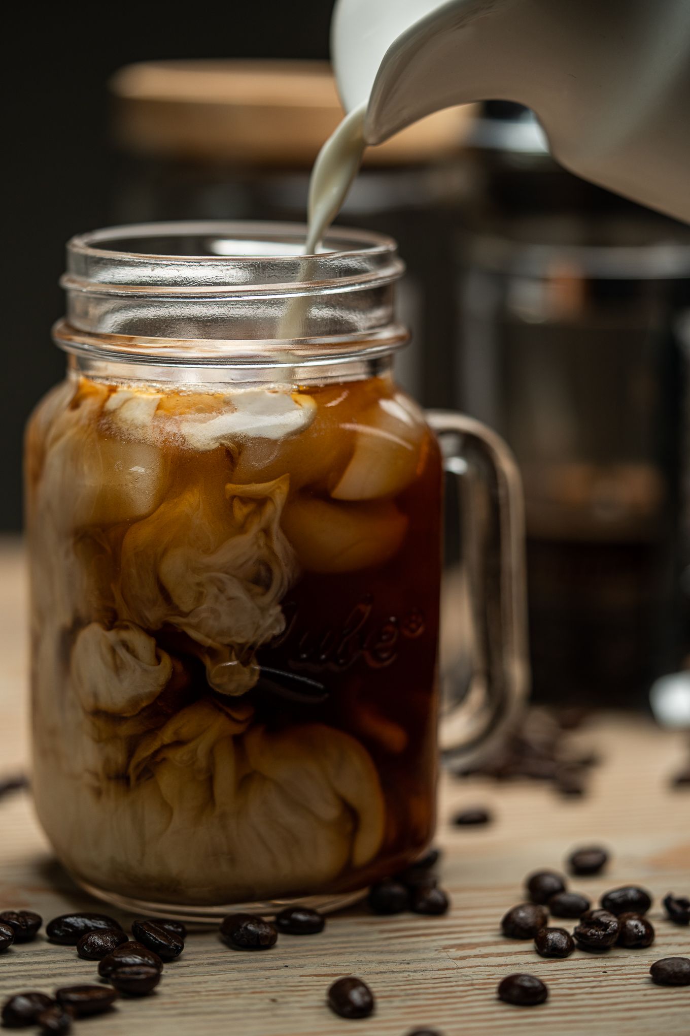 Cream being poured into iced coffee in a mason jar mug surrounded by coffee beans.