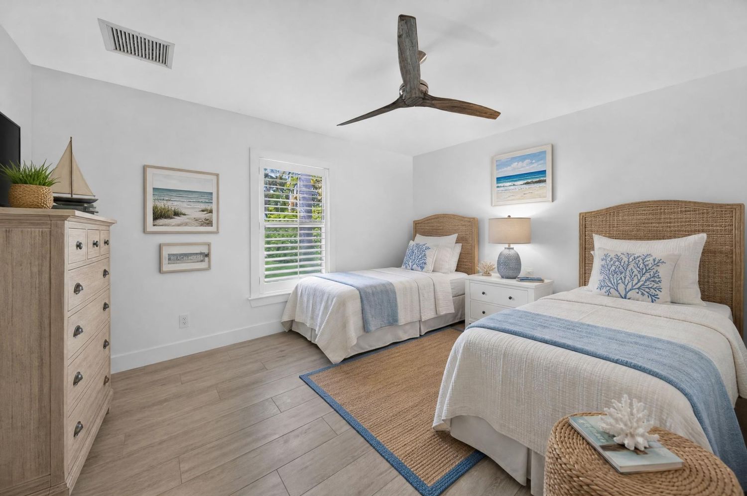 Bright coastal bedroom with twin beds, wicker headboards, ceiling fan, and beach-themed decor.