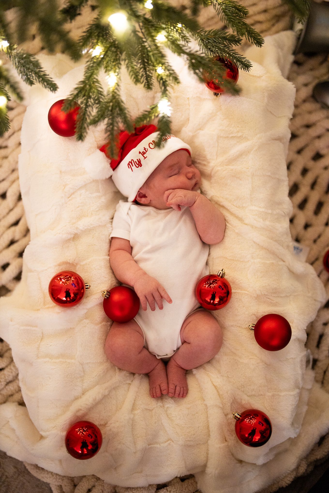 Newborn baby sleeping in a Santa hat surrounded by red Christmas ornaments under a festive tree.