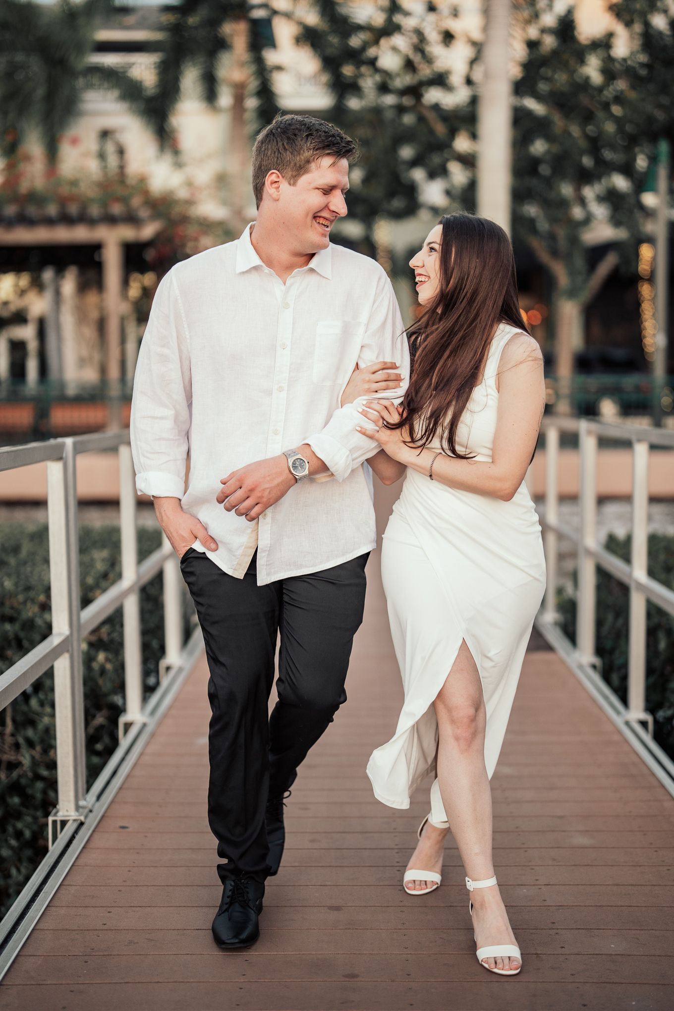 A smiling couple walks arm in arm on a wooden bridge, laughing and enjoying each others company.