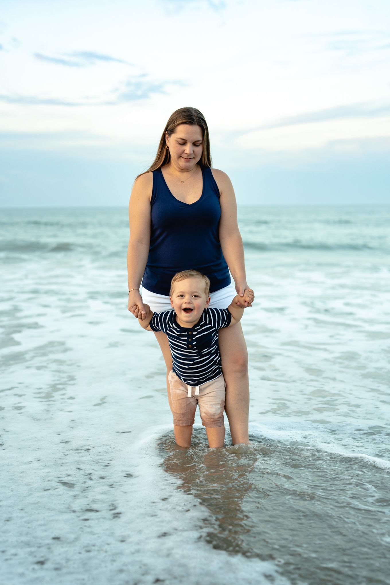 Mother standing in ocean waves holding toddler baby boy's hands on beach at dusk.