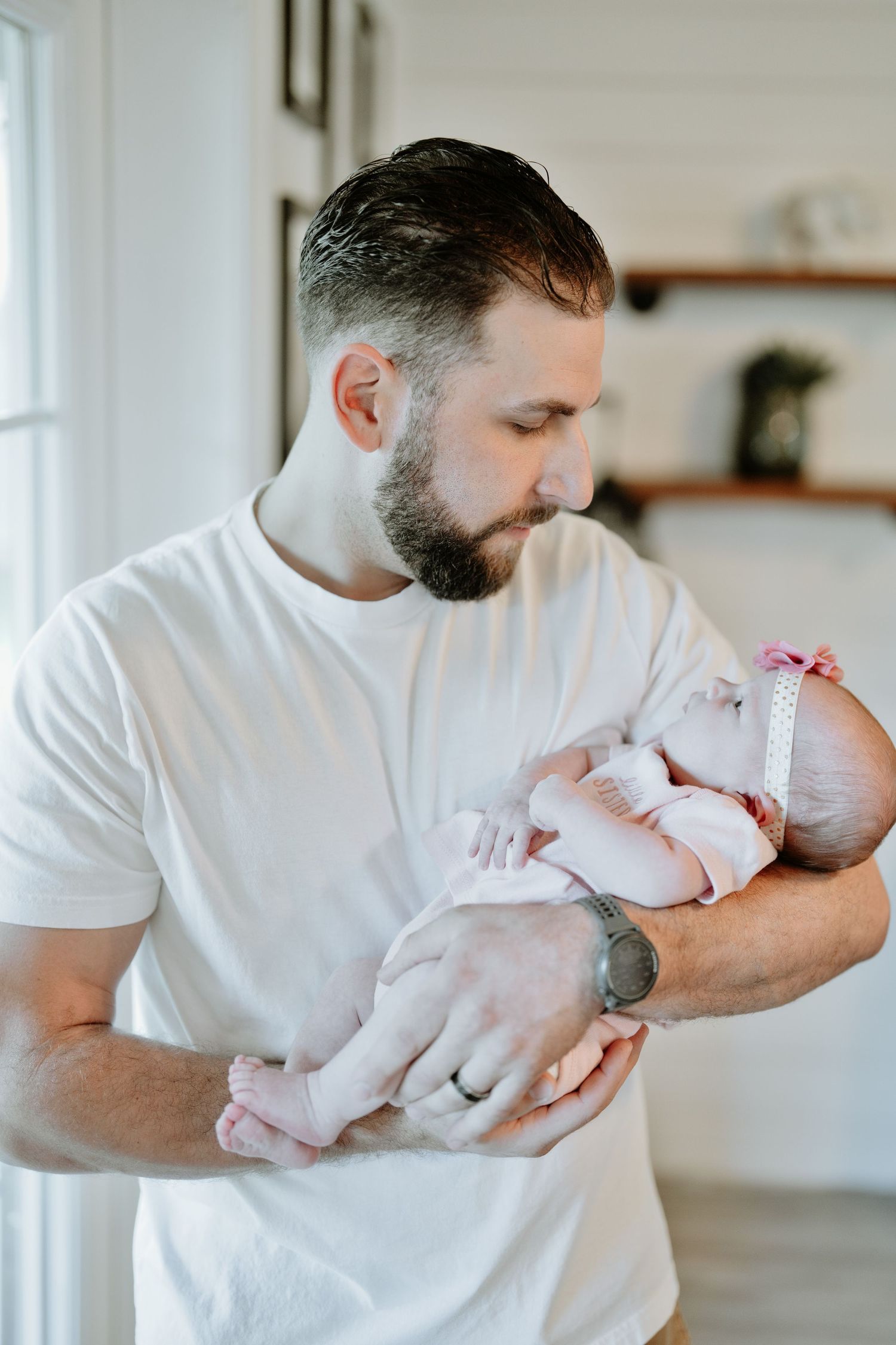 Bearded father in white shirt tenderly holding sleeping newborn baby girl in bright home.