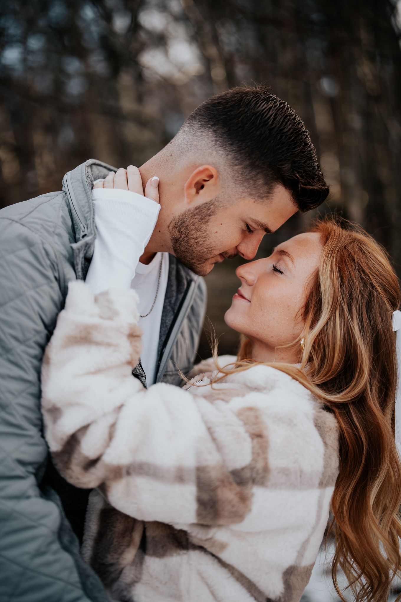 Couple sharing a romantic moment in a winter forest, foreheads touching, wearing cozy jackets.