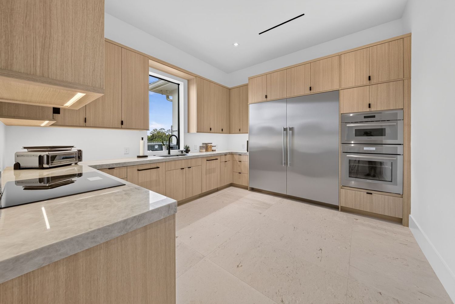 Contemporary kitchen featuring light wood cabinetry, stainless steel refrigerator, double ovens, and marble counters.
