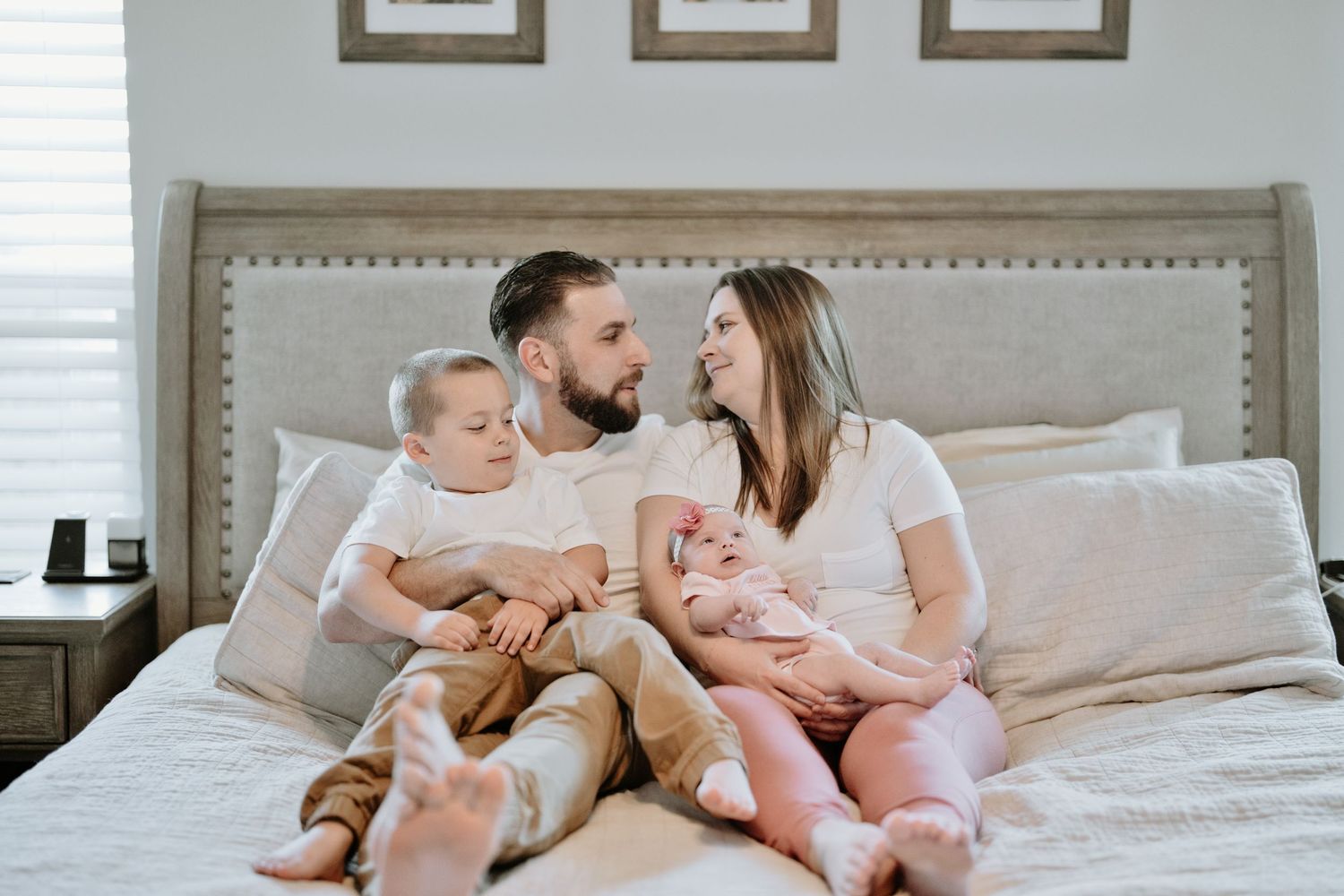 Happy family of four sitting on bed, parents gazing at each other with two young children.