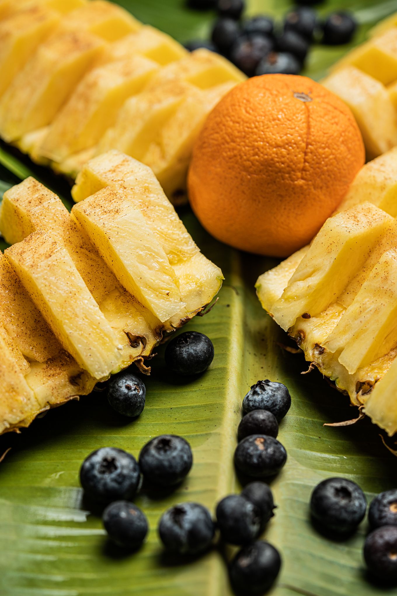 Fresh sliced pineapple, whole orange, and blueberries arranged on a tropical banana leaf.