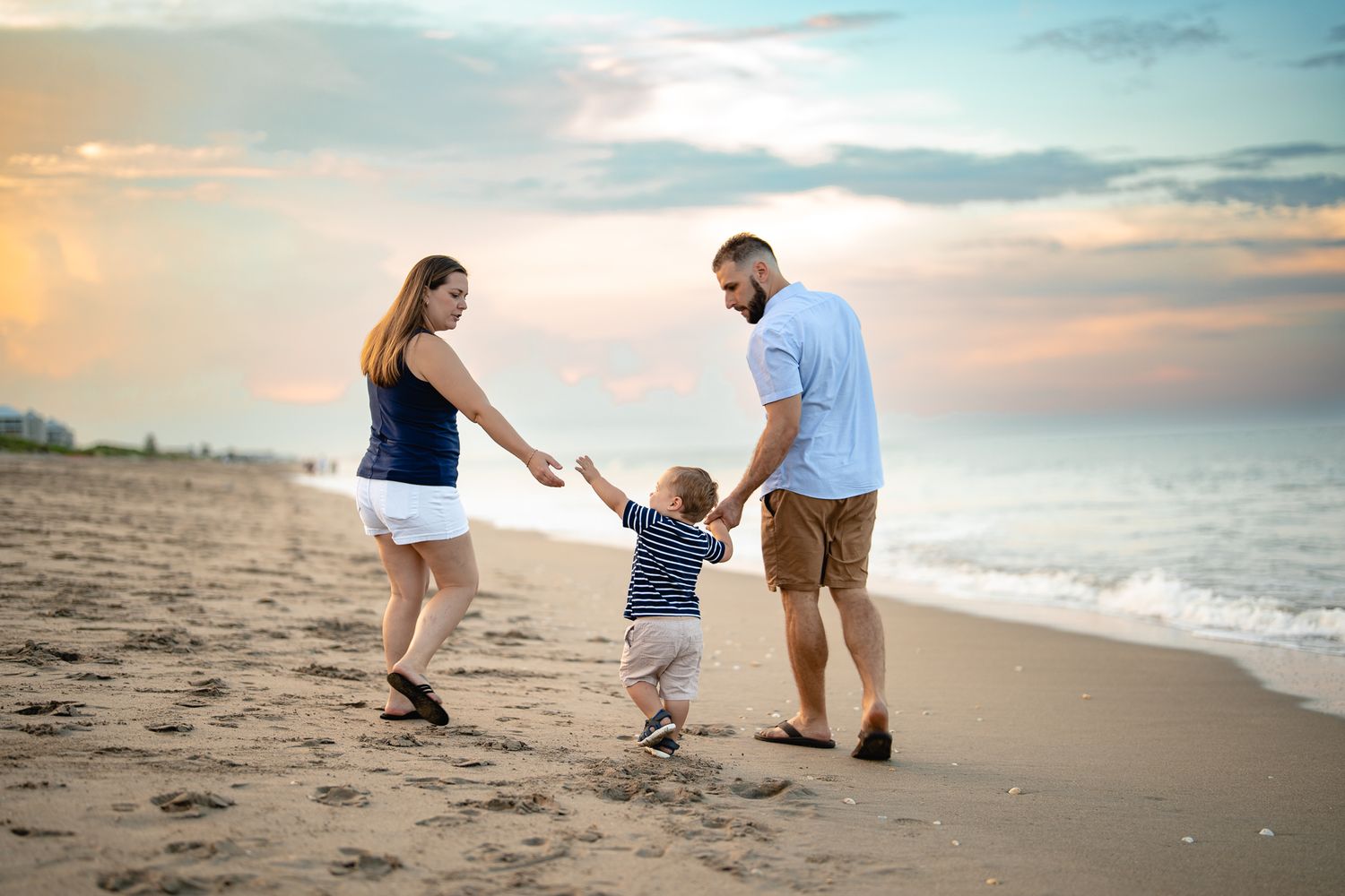 Happy family playing on sandy beach at sunset, parents swinging toddler son between them.