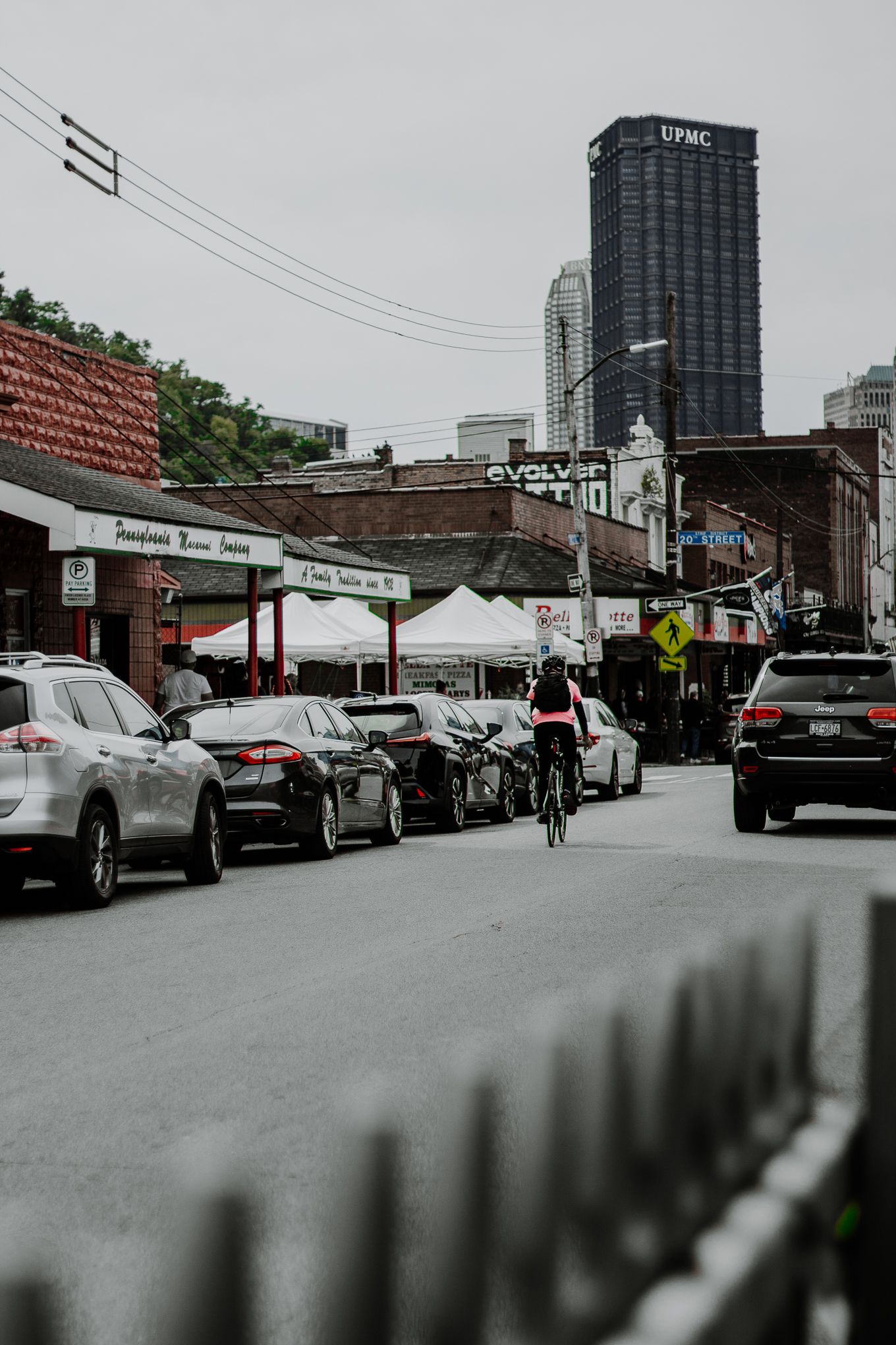 Busy urban street scene with parked cars, cyclist, and UPMC skyscraper in Pittsburgh city background.