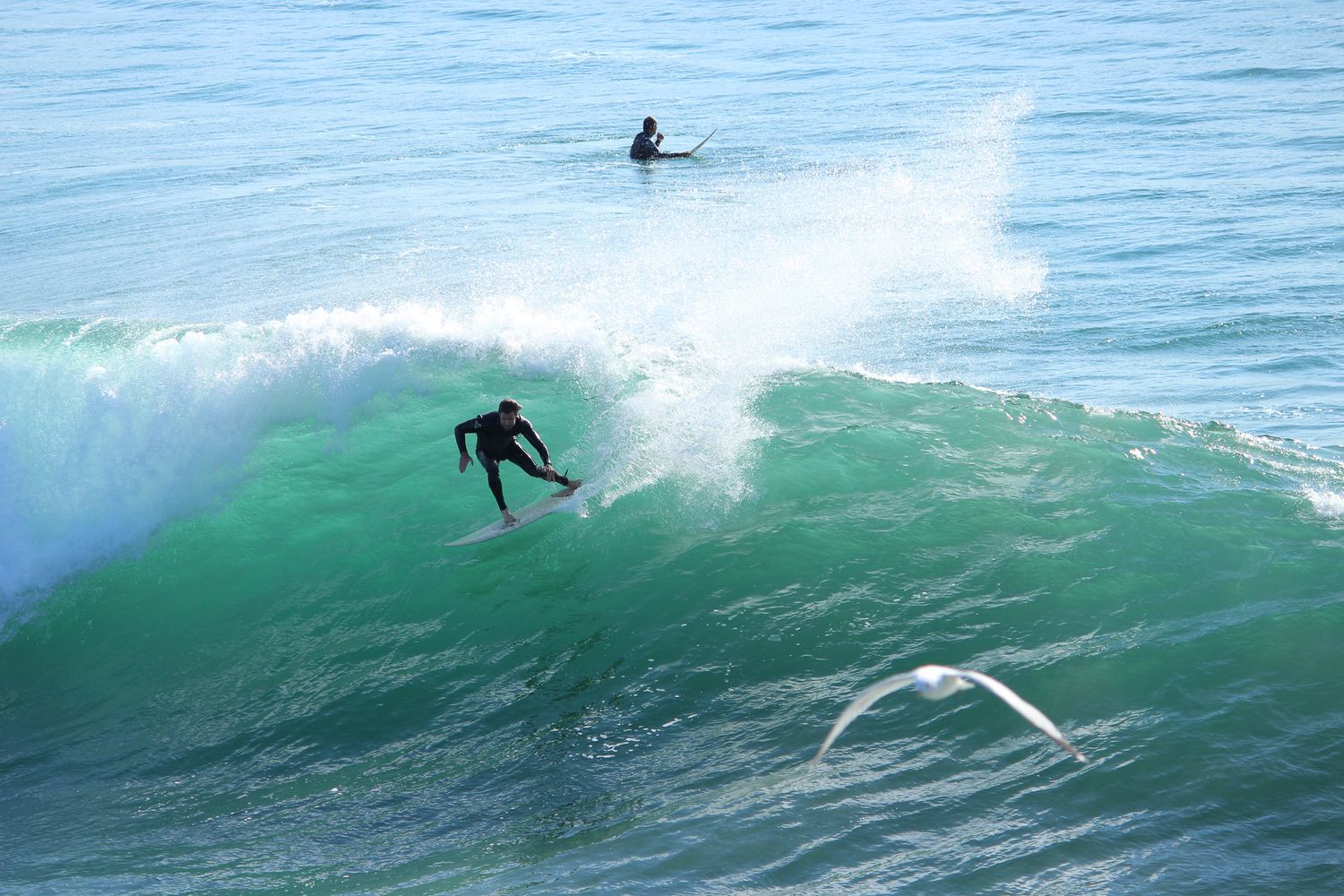 Surfer riding a large green wave in the ocean while another surfer waits in background.