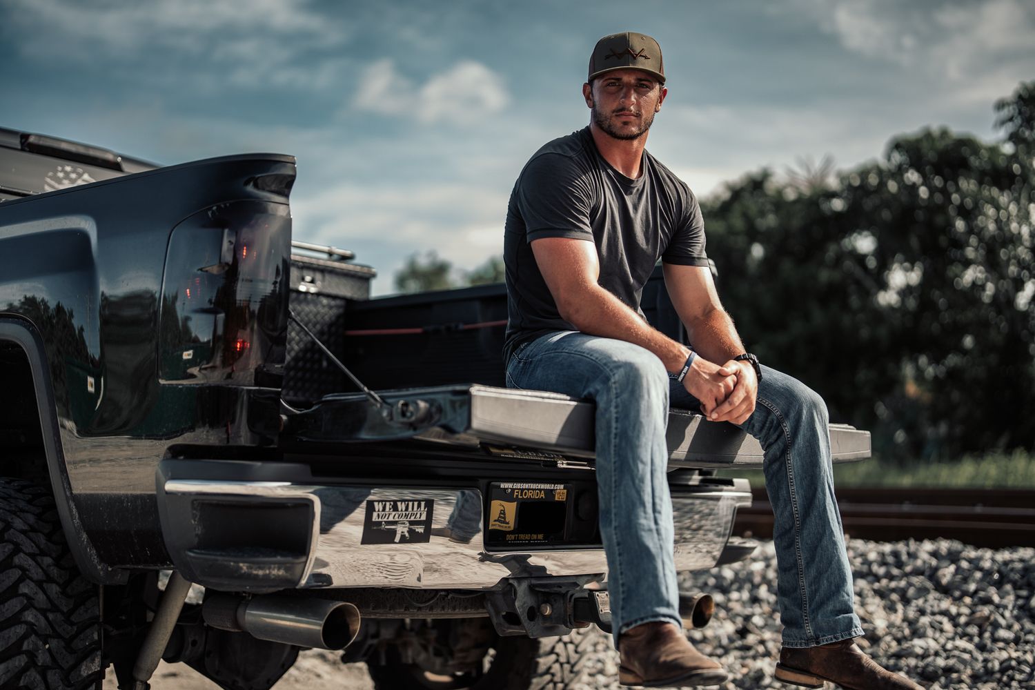 Rugged man in cap and jeans sitting on truck tailgate near railroad tracks under cloudy sky.