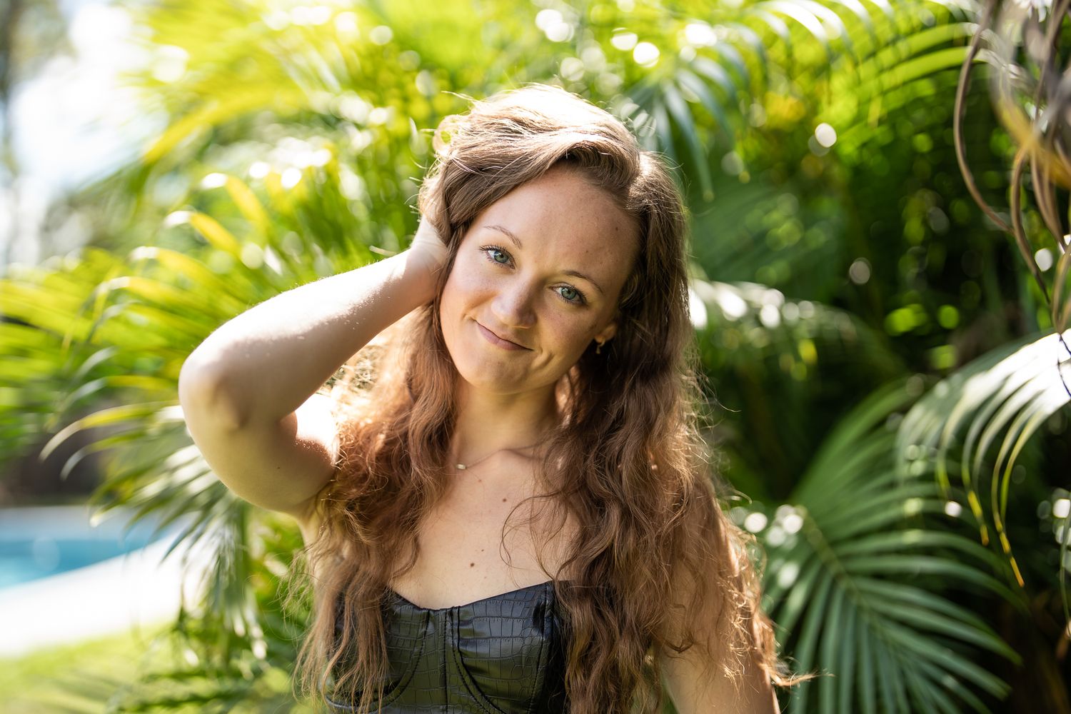 Smiling woman with long wavy hair posing outdoors among lush tropical palm leaves near a pool.