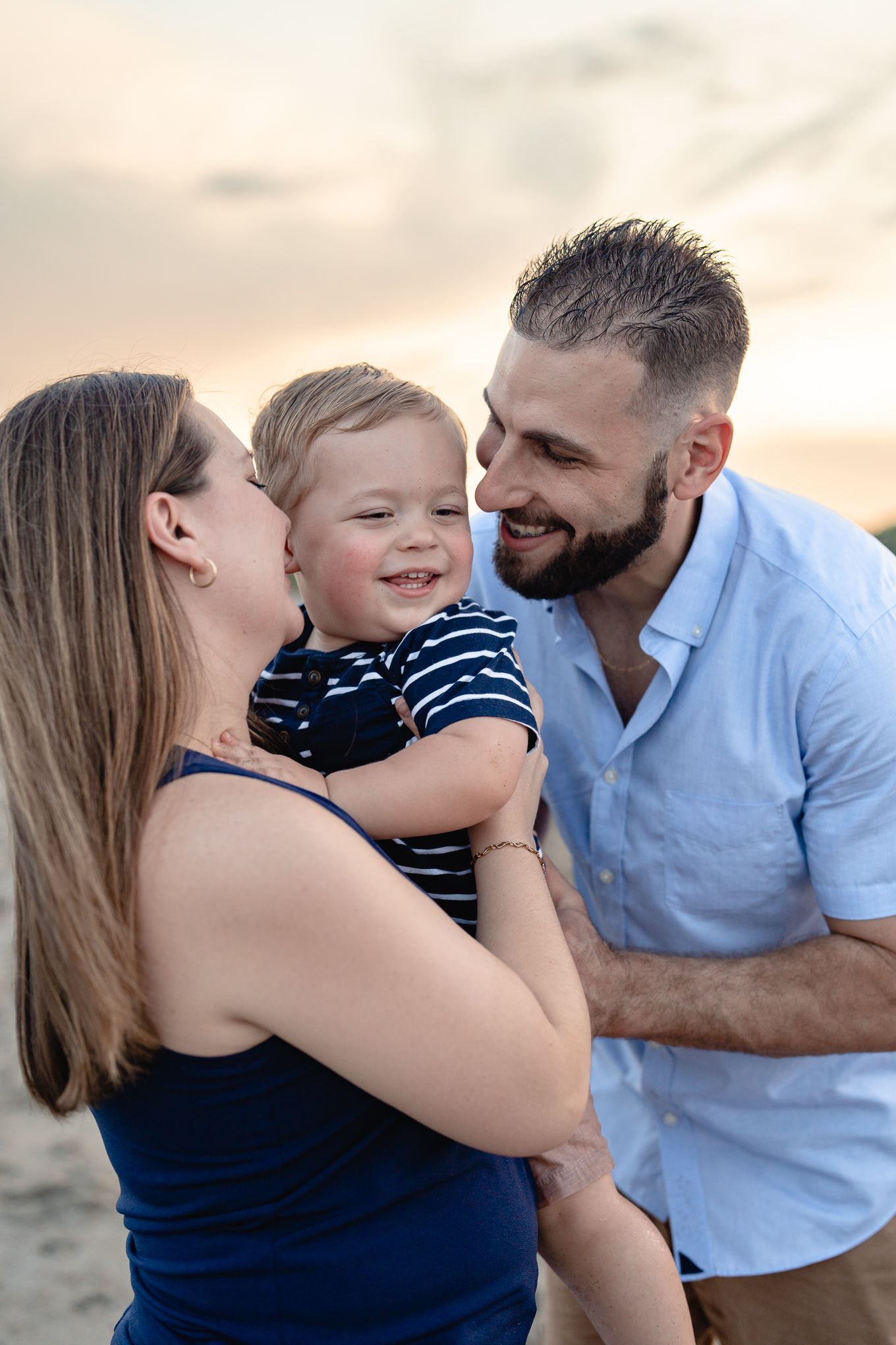 Loving parents smiling at happy laughing toddler boy on beach during colorful sunset.