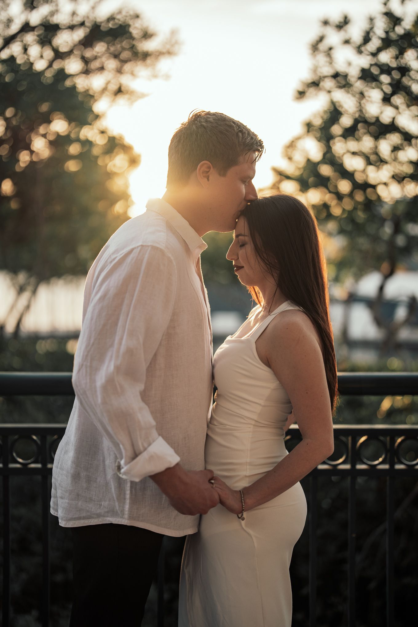Romantic couple embracing at sunset near a decorative railing with glowing backlight behind them.