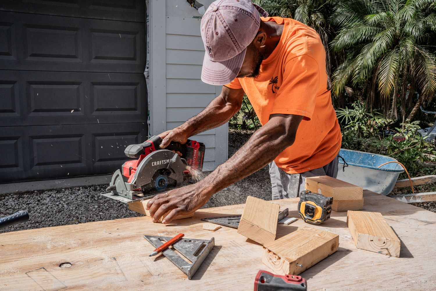 Construction worker in orange shirt using circular saw to cut lumber on outdoor workbench.