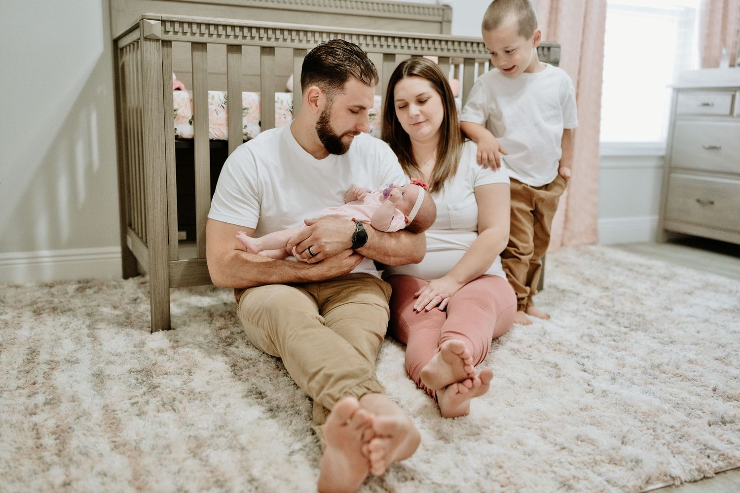 Young family with newborn baby and toddler sitting on nursery floor by wooden crib.