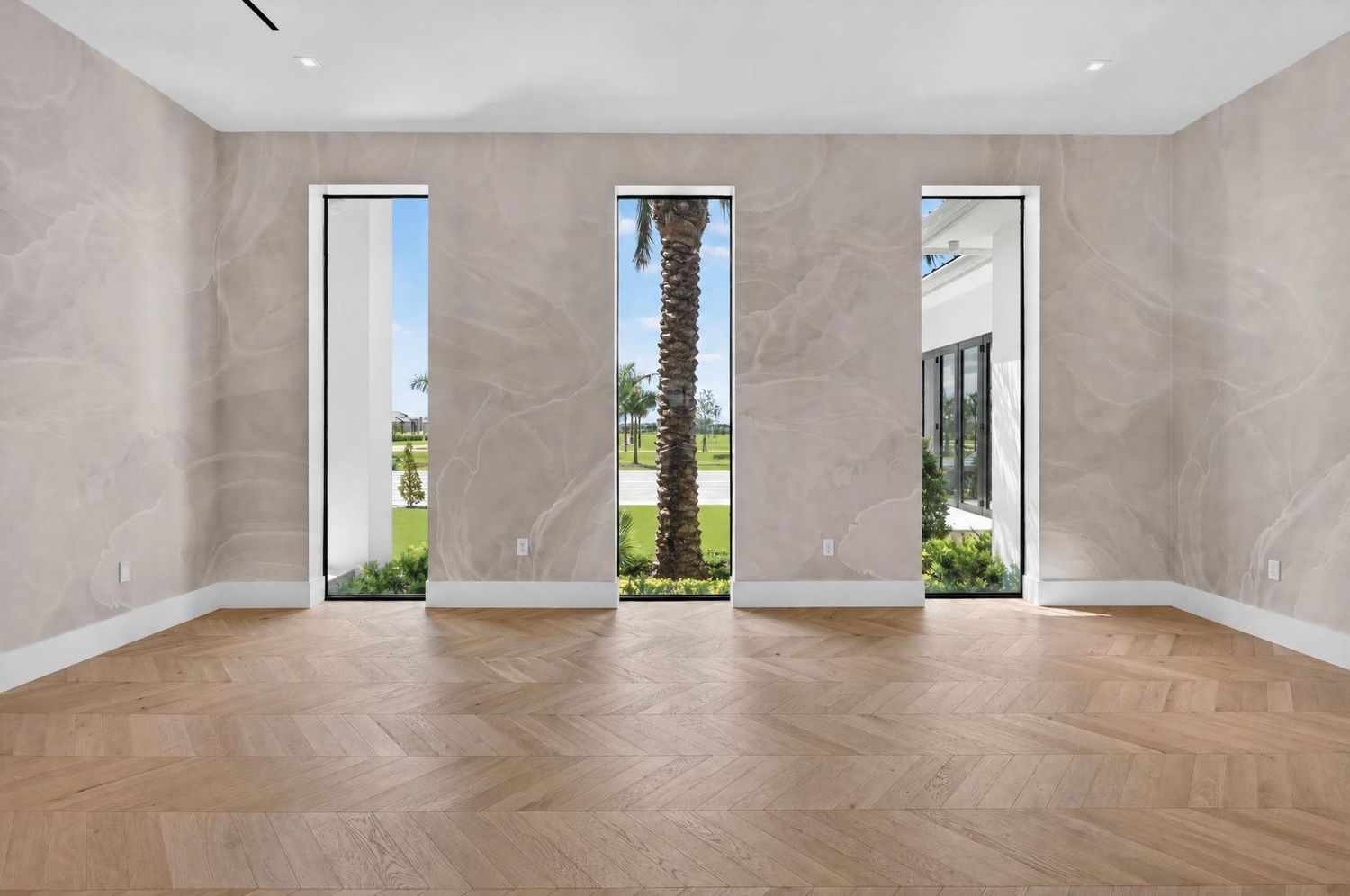 Luxurious entryway with herringbone wood floors, marble walls, tall windows, and view of palm tree outside.