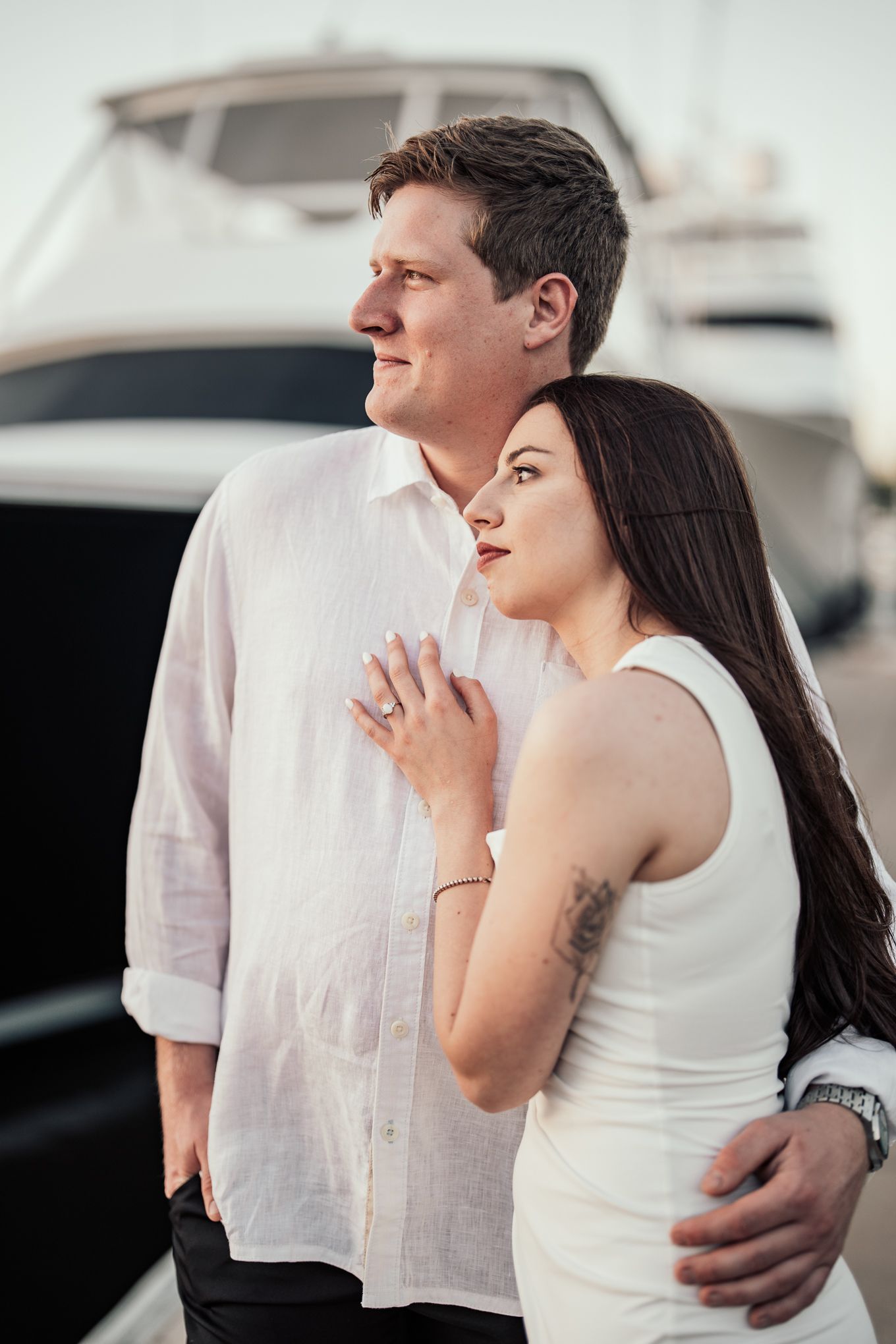 Couple embracing near luxury yacht marina, woman in white dress resting head on mans shoulder.