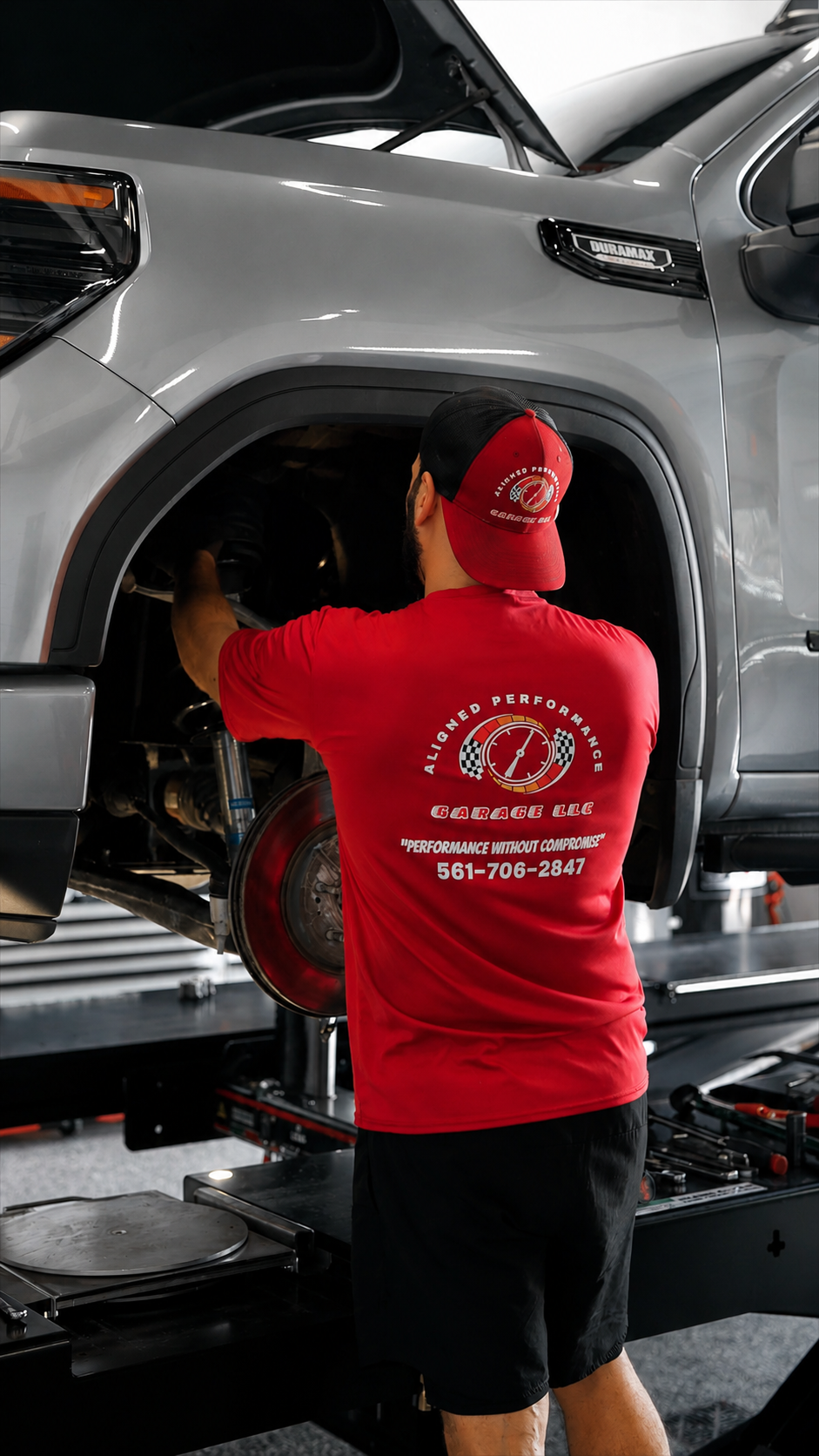 Aligned Performance mechanic in red uniform performing wheel alignment on lifted silver SUV.