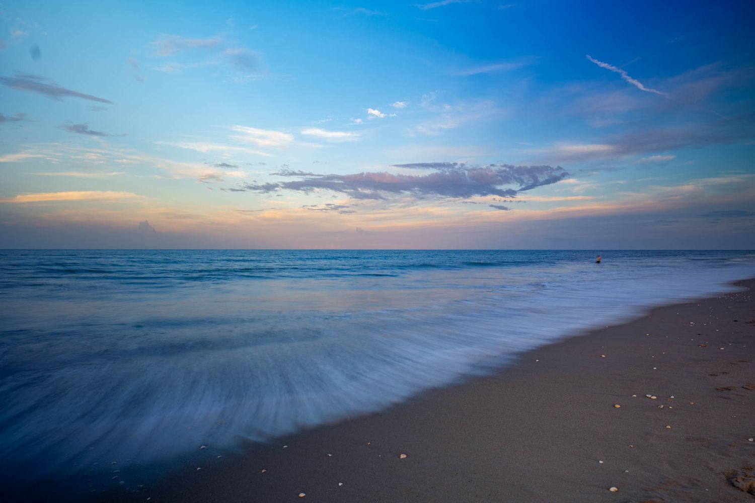 Long exposure shot of ocean waves washing onto sandy beach at sunset with colorful sky.