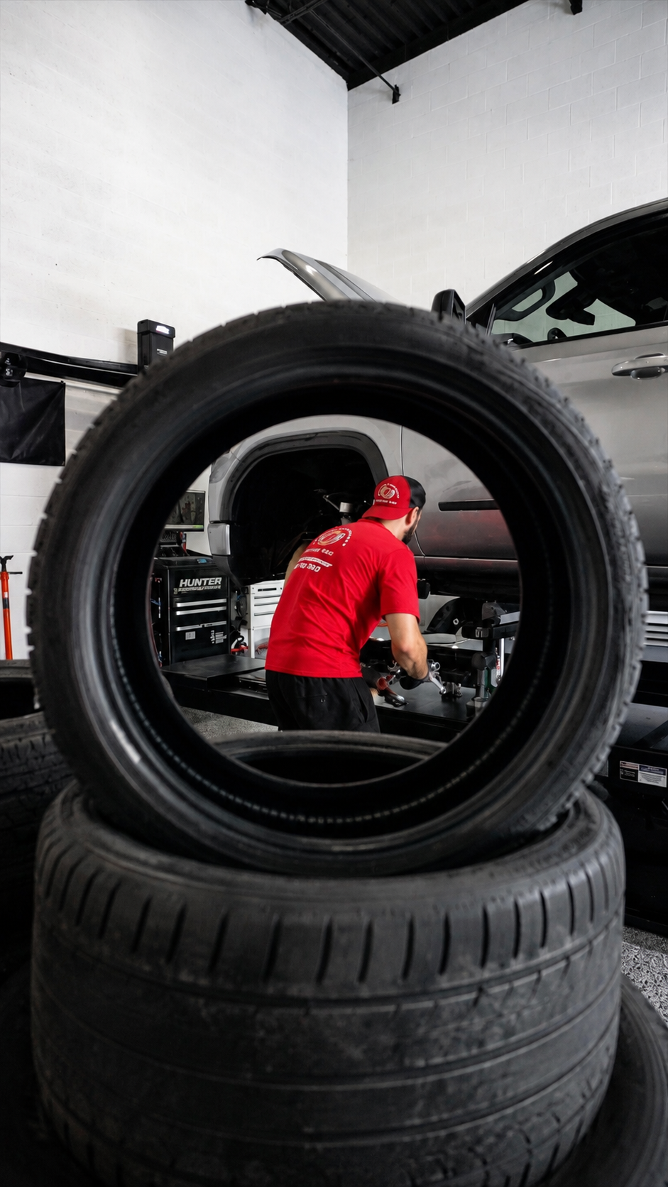 Auto technician in red Aligned Performance shirt working on car viewed through stacked tires.