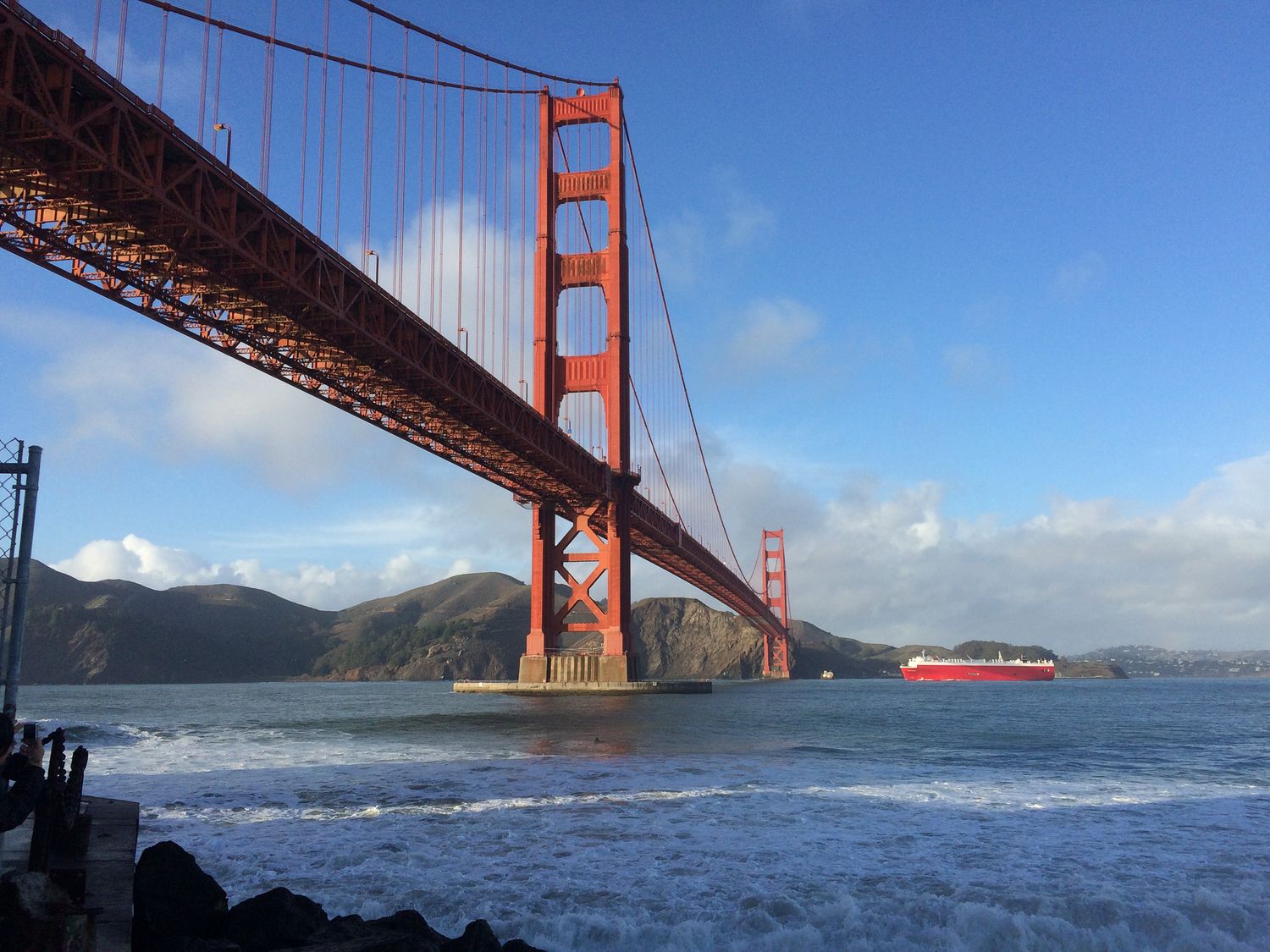 Golden Gate Bridge spanning San Francisco Bay with a red cargo ship passing beneath on a sunny day.
