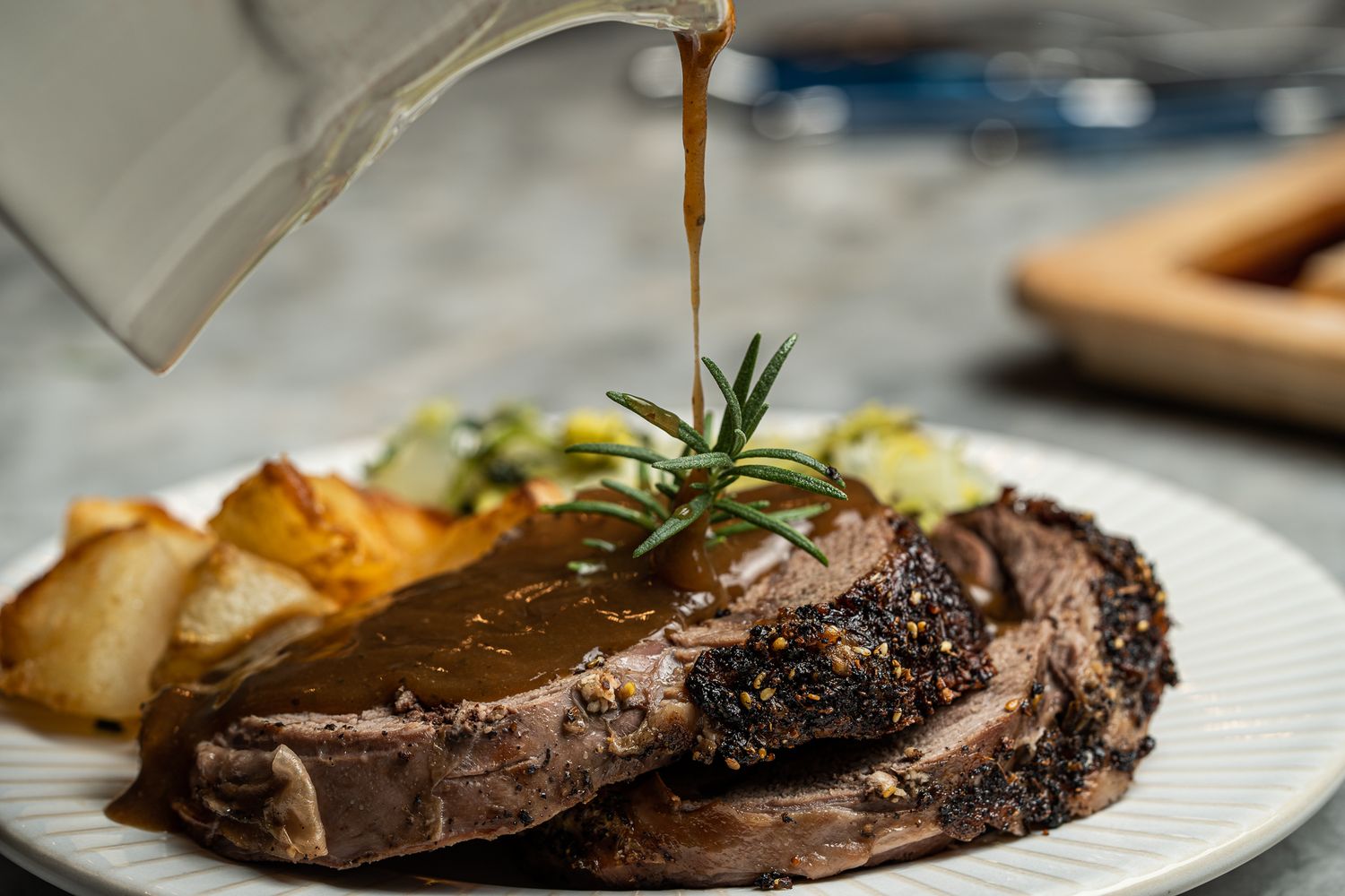 Gravy being poured over sliced roast beef with roasted potatoes and rosemary garnish.