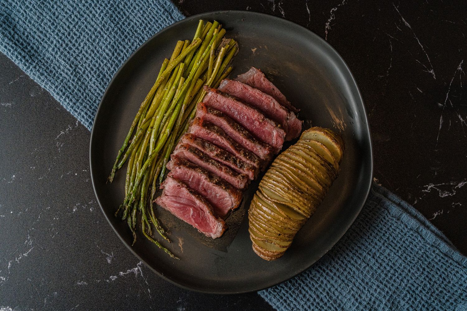 Overhead view of sliced medium-rare steak served with asparagus and hasselback potato on dark plate.