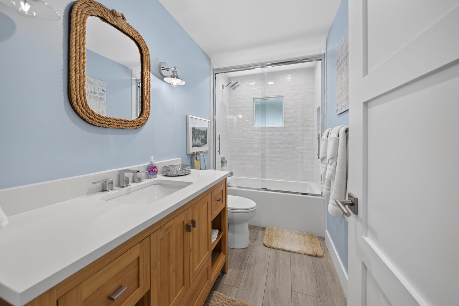 Light blue bathroom with wood vanity, rope mirror, and combined shower-tub with glass doors.
