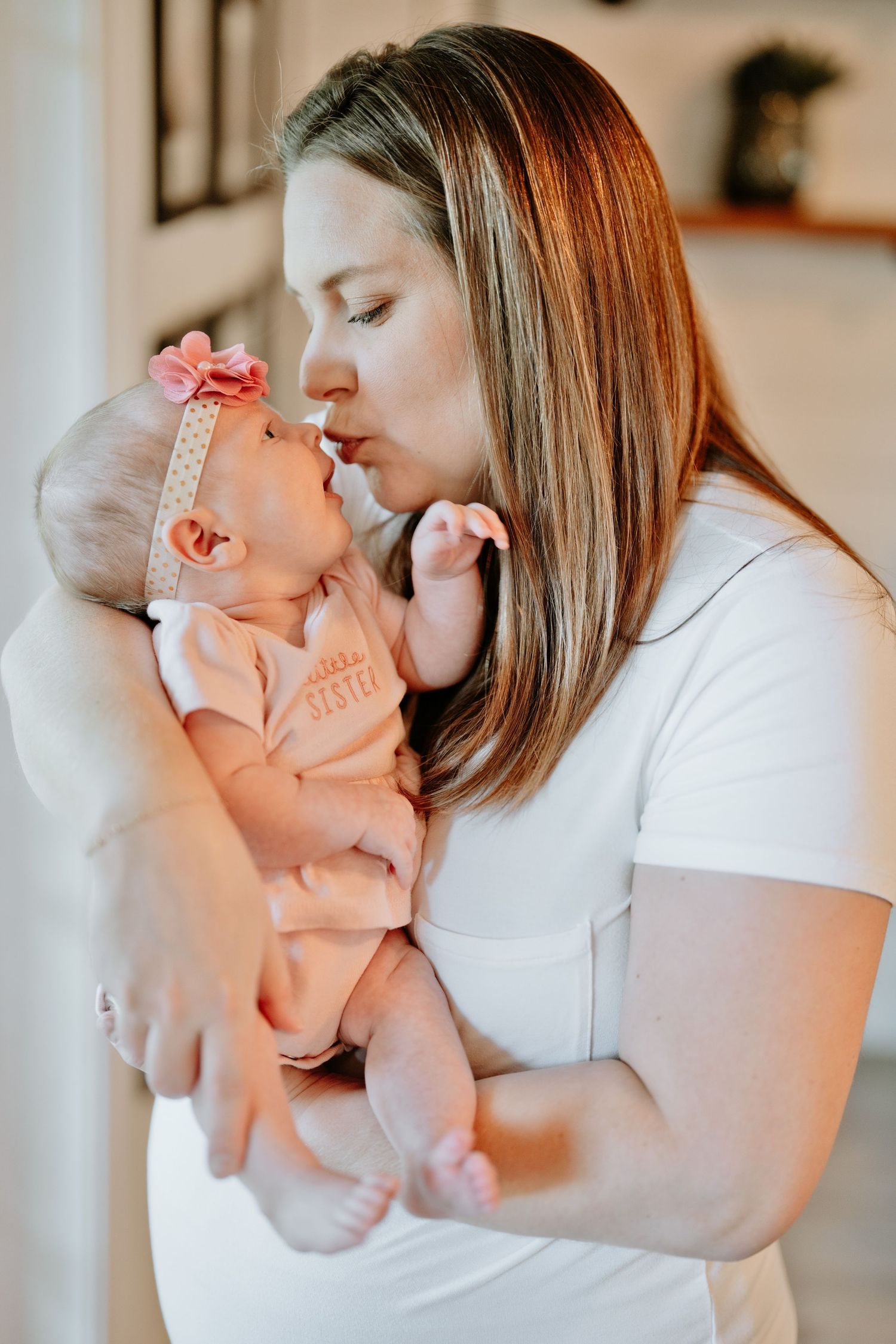 Mother kissing newborn baby girl wearing pink bow headband and little sister onesie indoors.