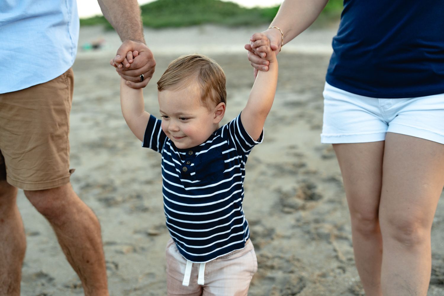 Close-up of toddler boy in striped shirt holding parents' hands while walking on sandy beach.