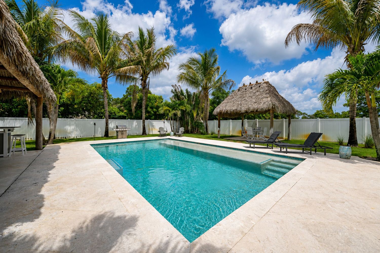 Private backyard oasis featuring a rectangular pool, thatched tiki hut, and tropical palm trees on a sunny day.