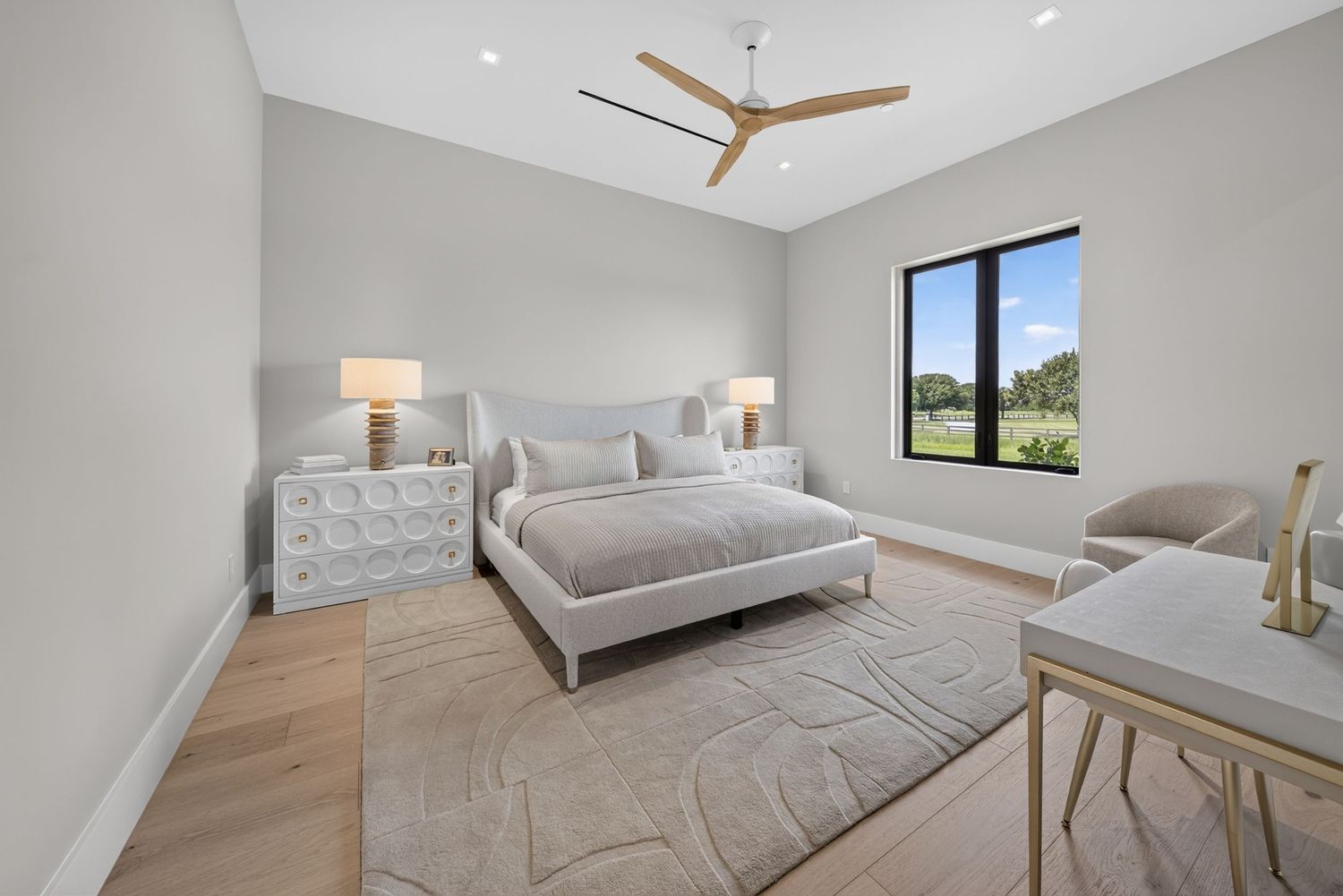 Bright bedroom with neutral decor, ceiling fan, area rug, white dresser, and large window overlooking greenery.