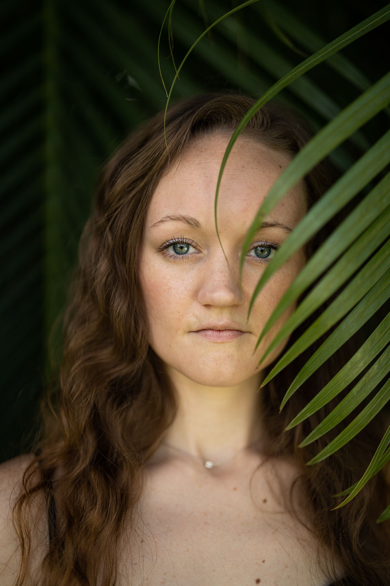 Young woman with freckles and blue eyes partially hidden behind tropical palm leaf in nature portrait.