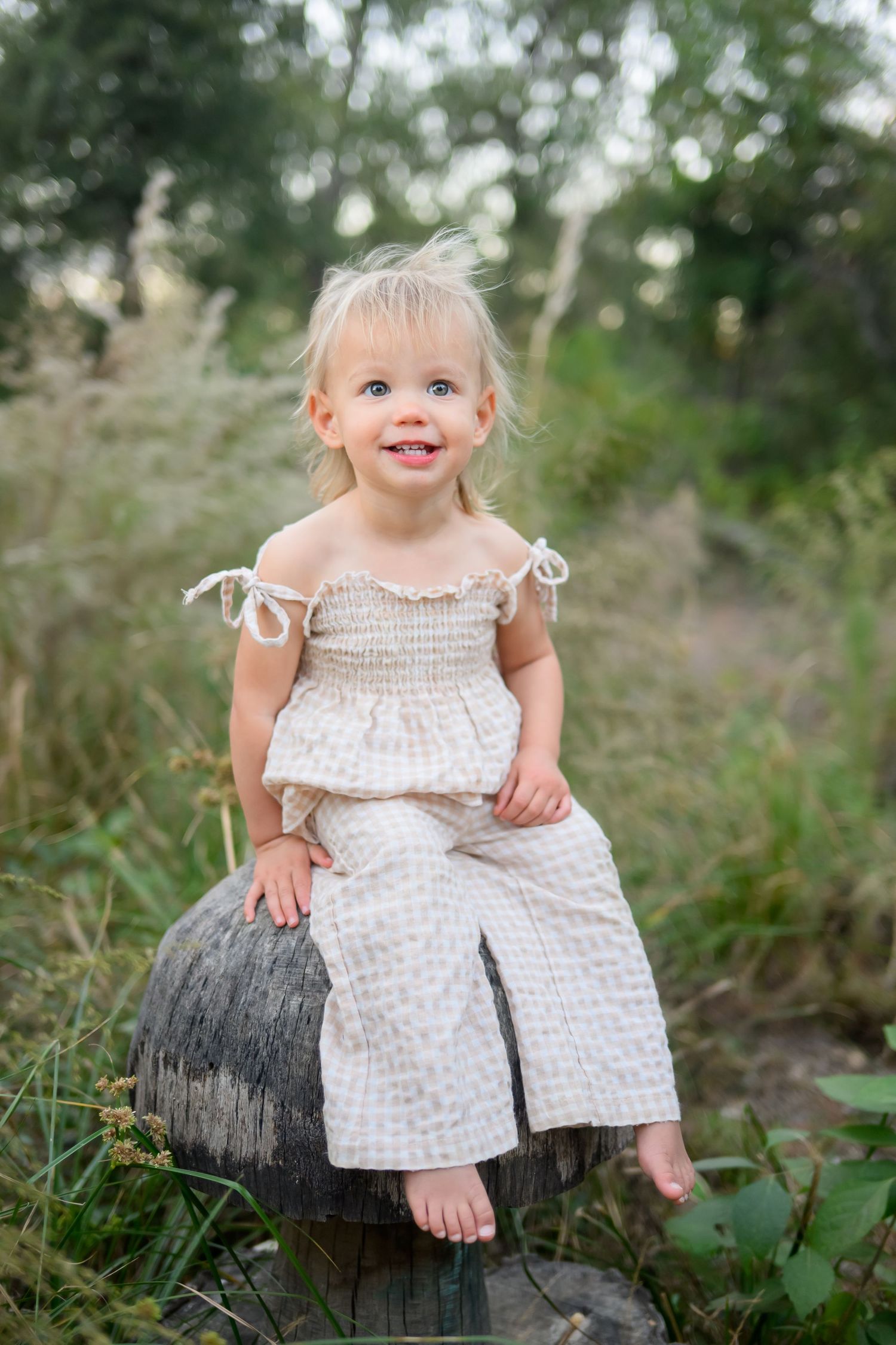 Smiling toddler girl in white smocked outfit sitting on a tree stump outdoors in a lush green field.