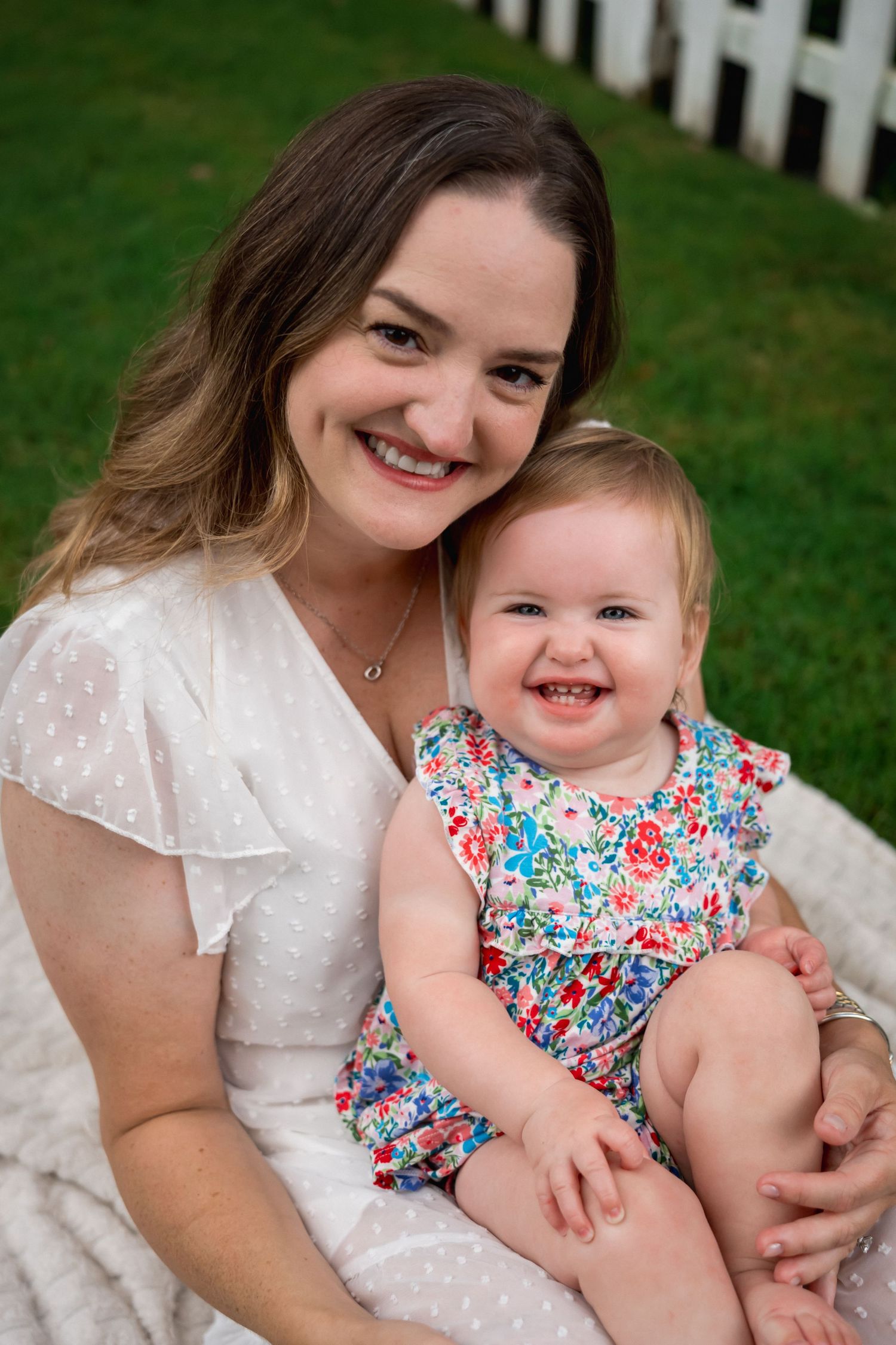 Smiling mother in white dress holding laughing baby girl in floral outfit on green lawn.