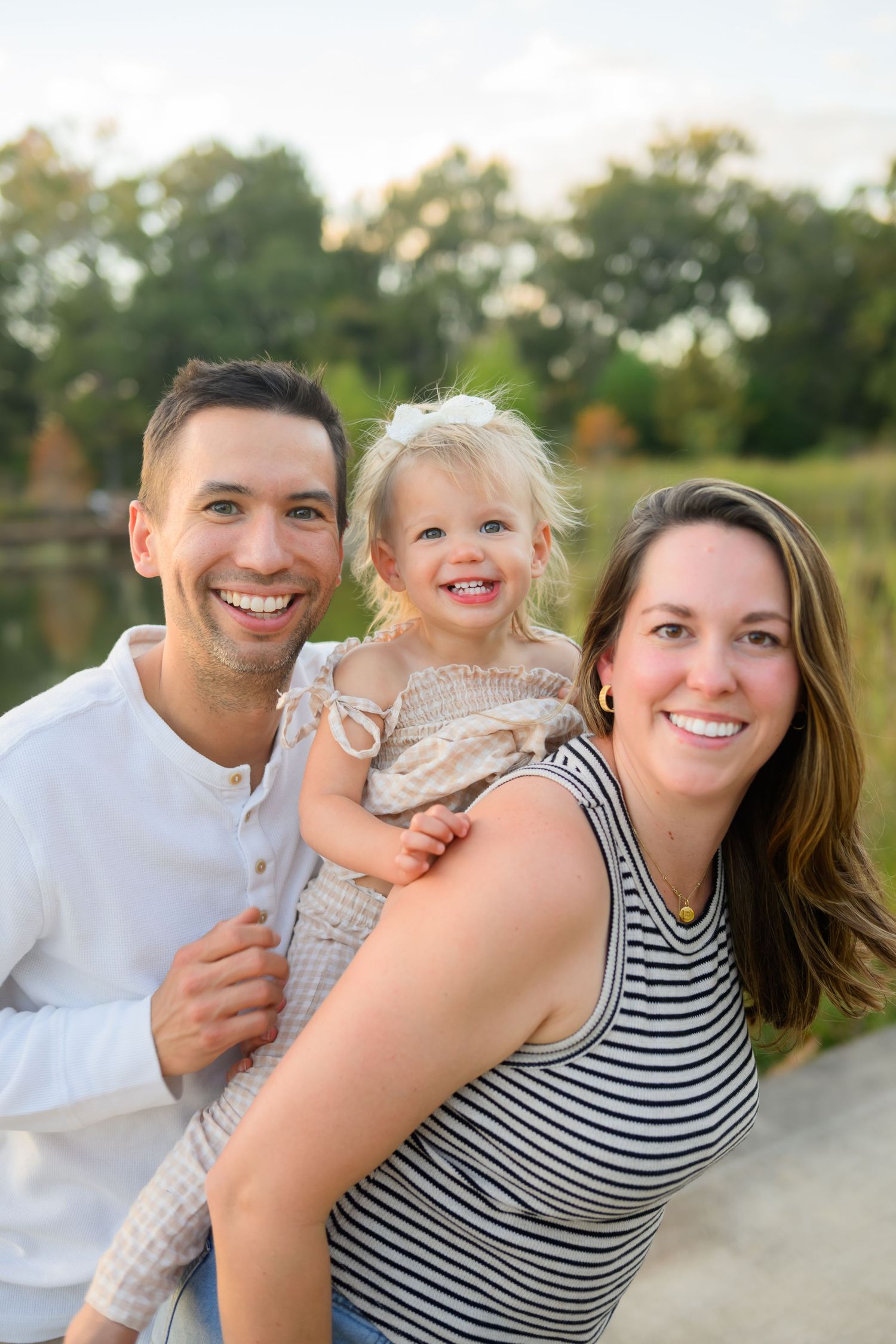 Happy family portrait outdoors with smiling parents holding their young blonde toddler daughter near a pond.