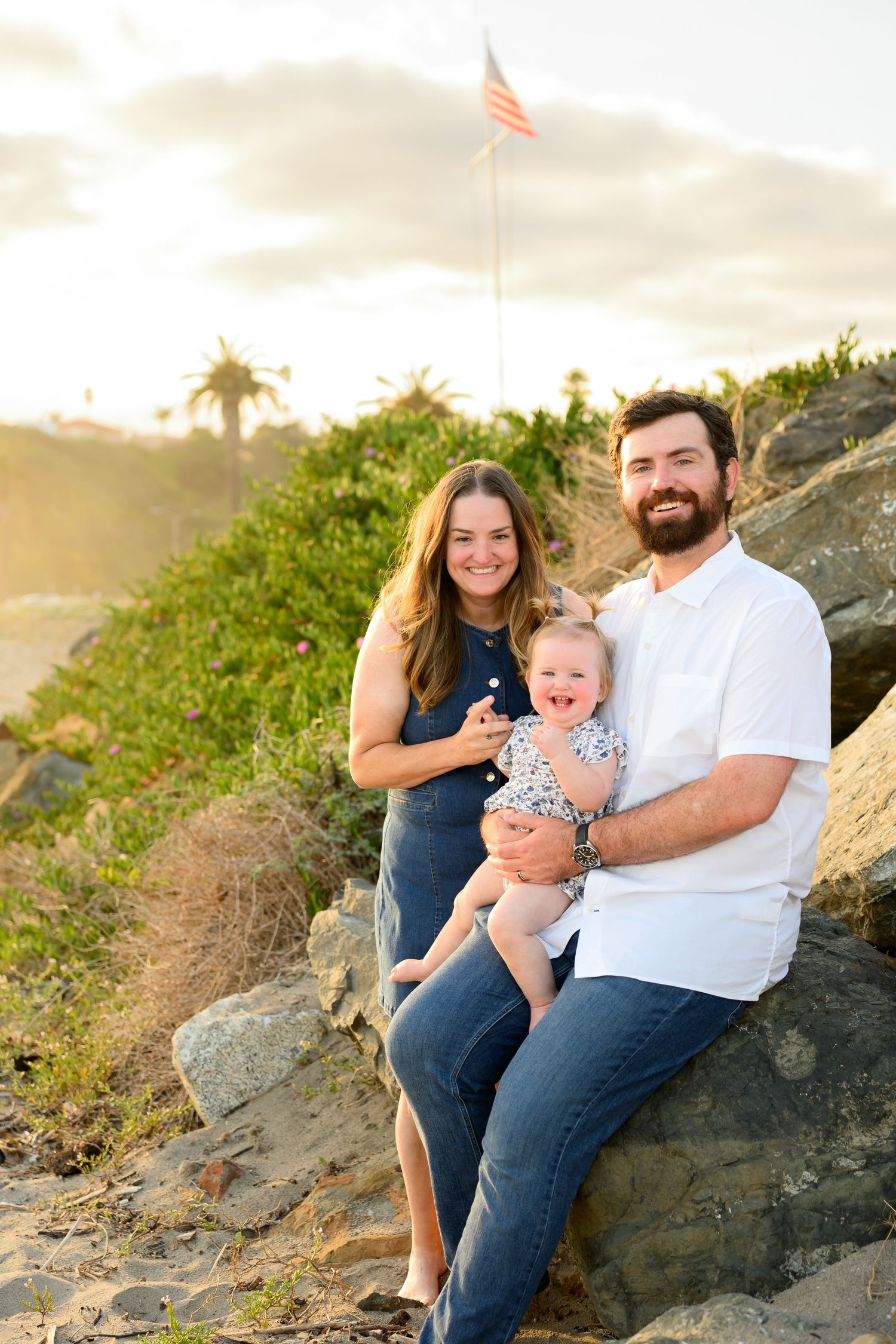 Smiling family with baby sitting on coastal rocks at sunset with American flag in background.