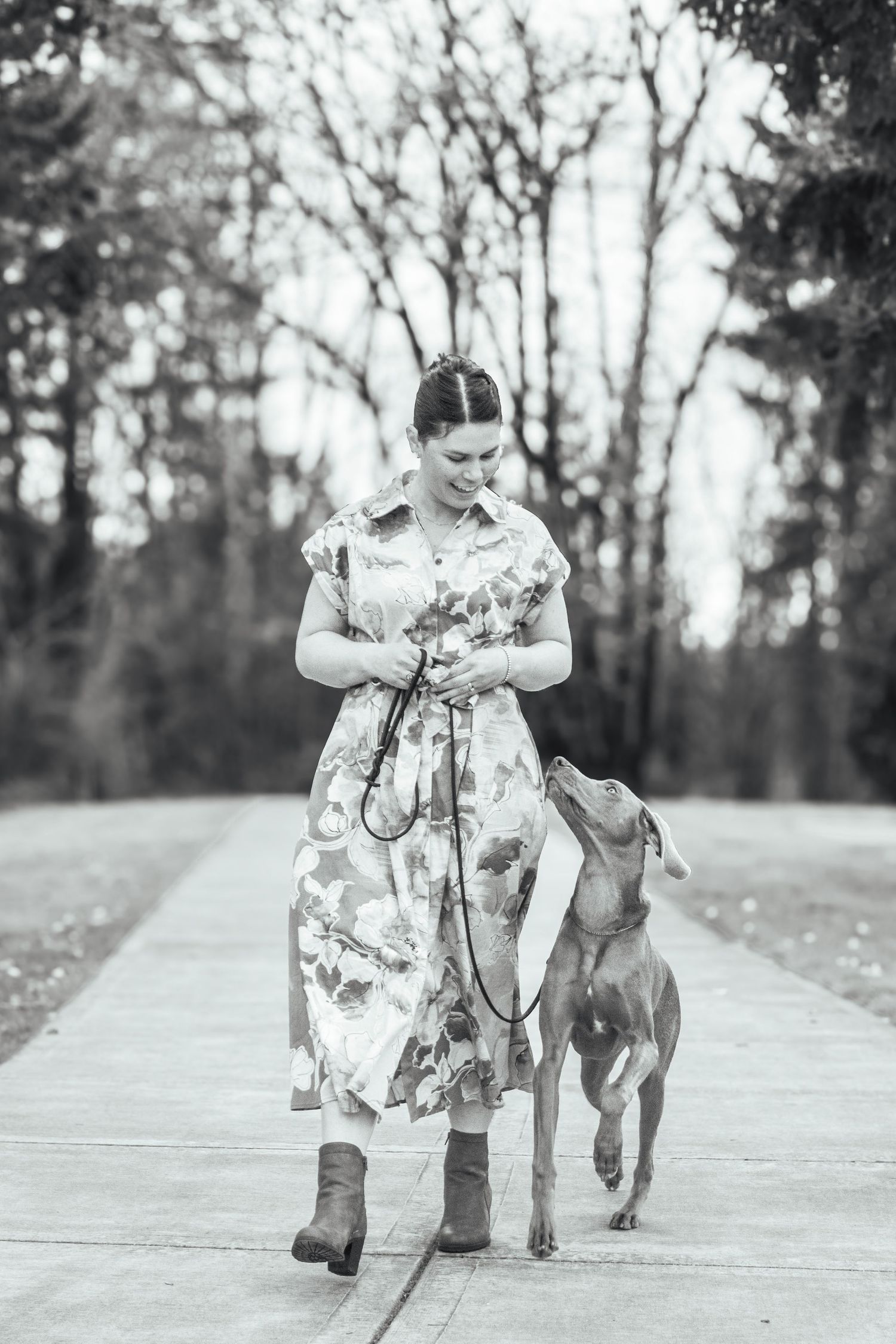 Black and white portrait of a woman walking with her dog along a park pathway.