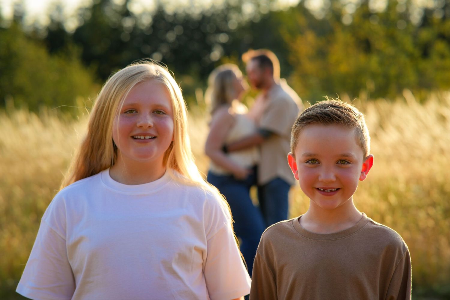 Two smiling siblings stand together in a sunlit field while their parents embrace in the softly blurred background during a family session