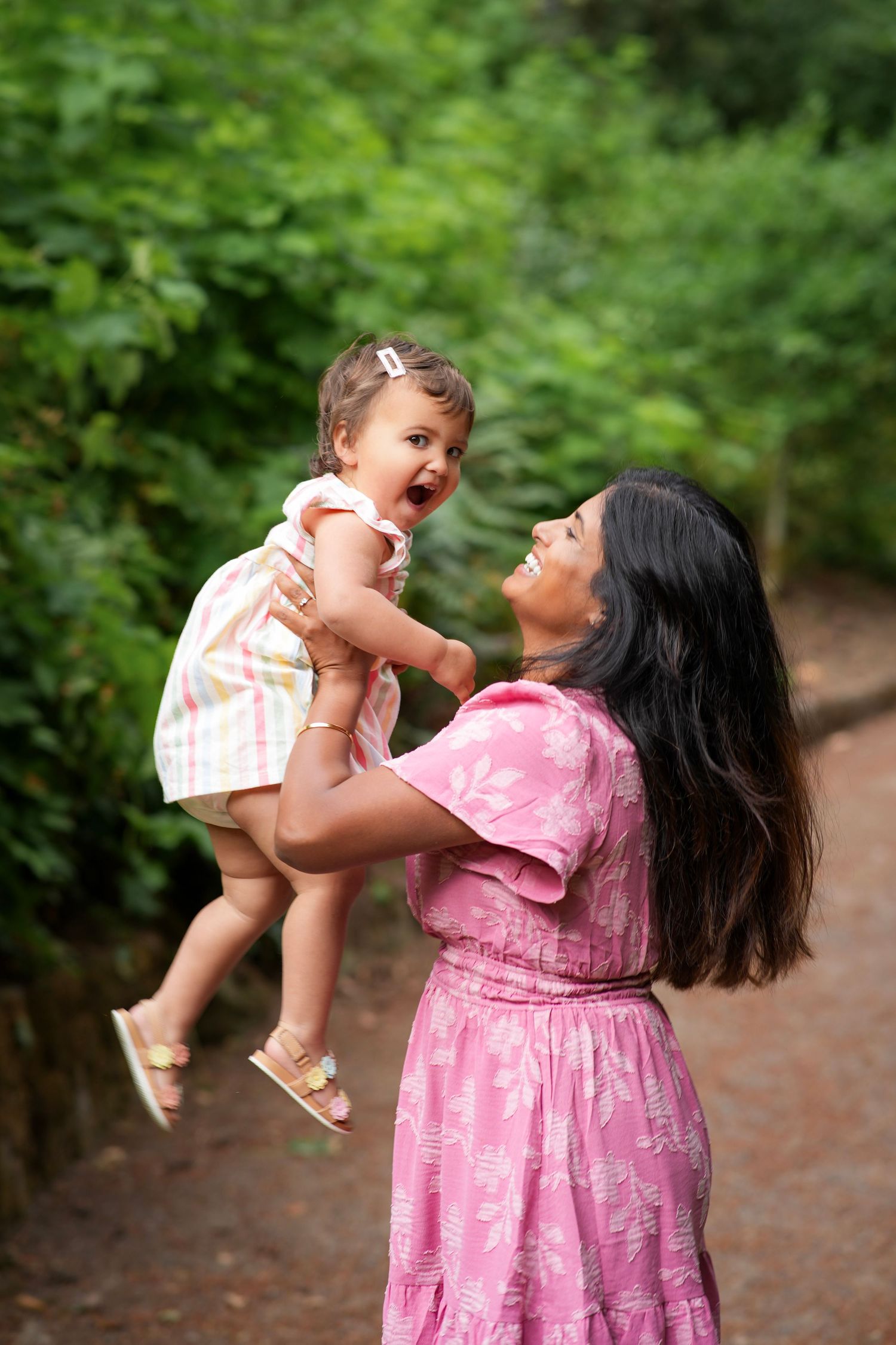 Mom laughing as she lifts her toddler daughter into the air during a playful outdoor family photography session