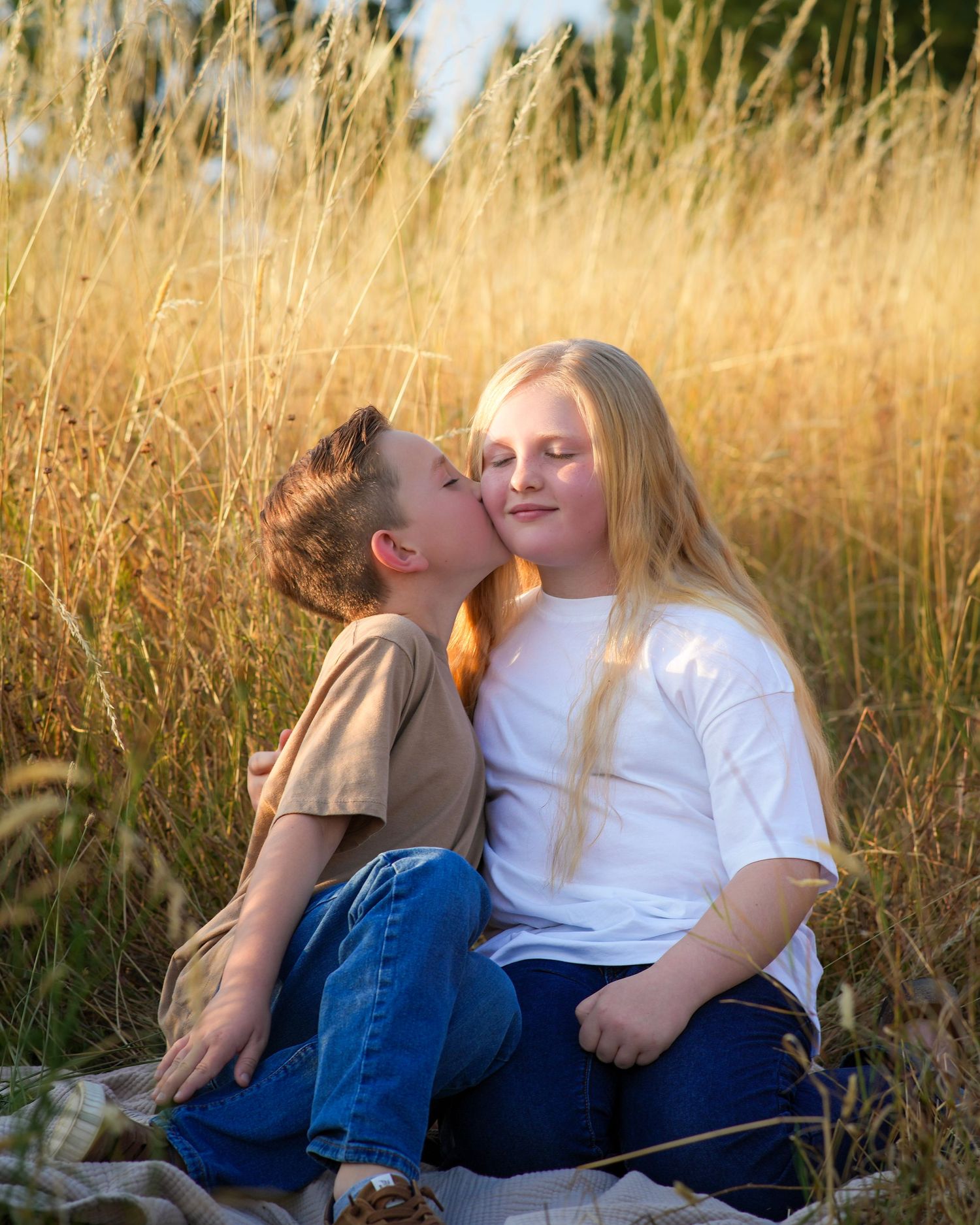 Young brother kissing his older sister on the cheek while they cuddle together in a golden field