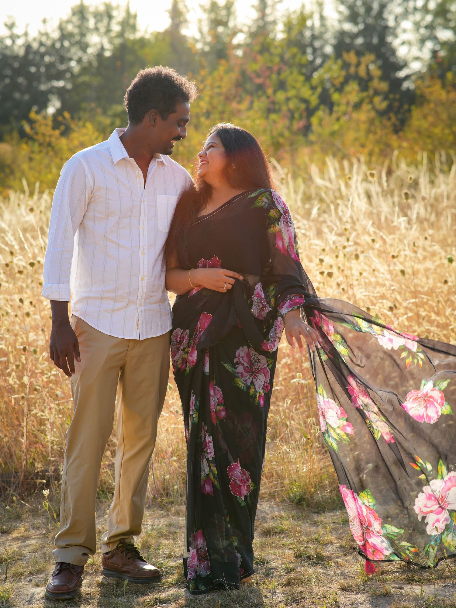 Couple smiling at each other in a golden field as her floral dress catches the breeze during a sunset session