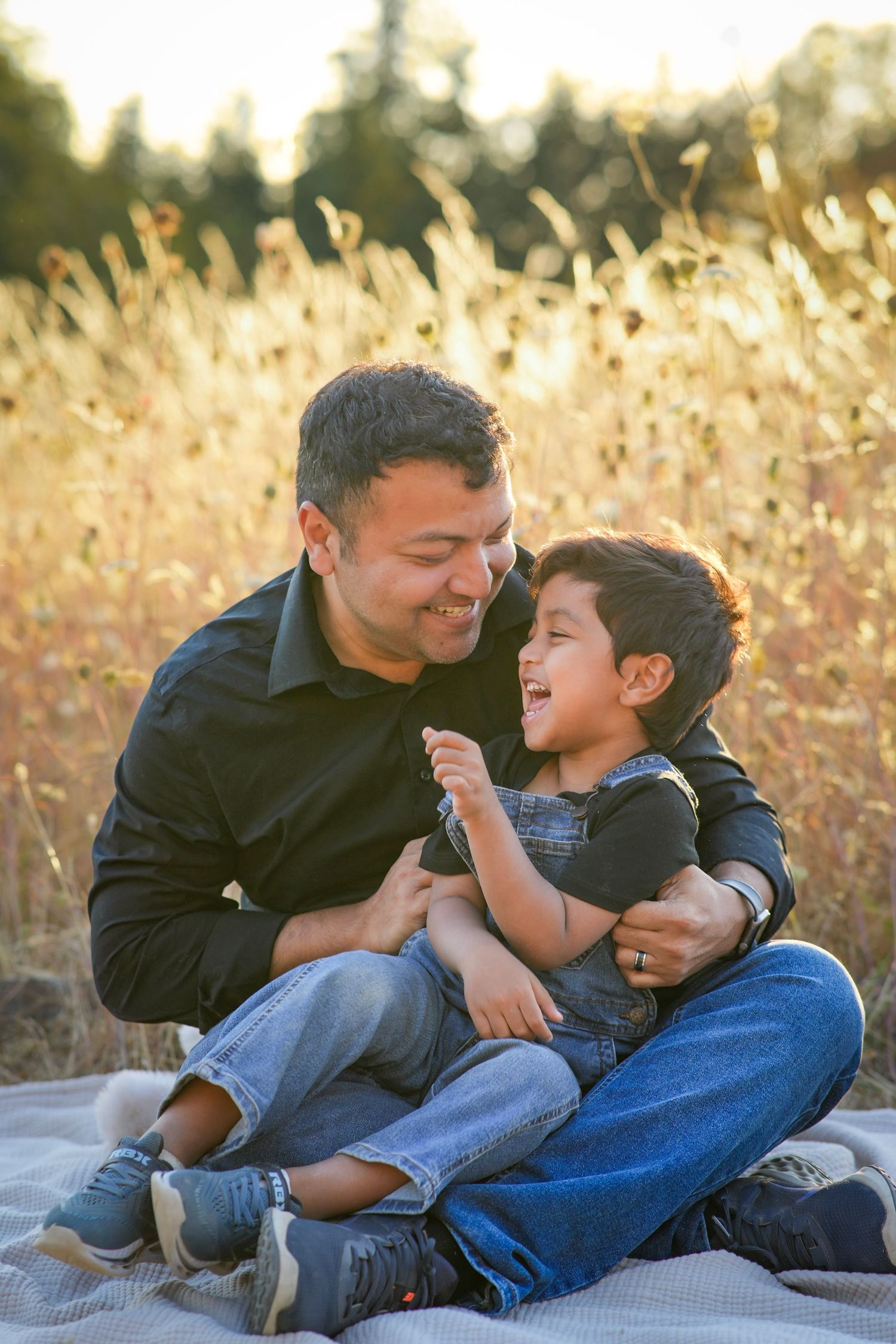 Dad and young son laughing together on a blanket during a candid outdoor family photography session