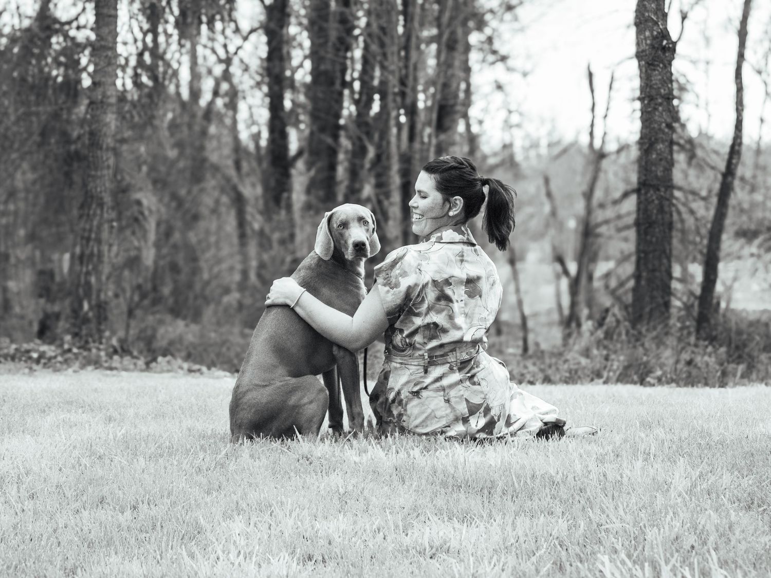 Black-and-white portrait of a woman sitting in the grass and embracing her dog during an outdoor pet photography session.