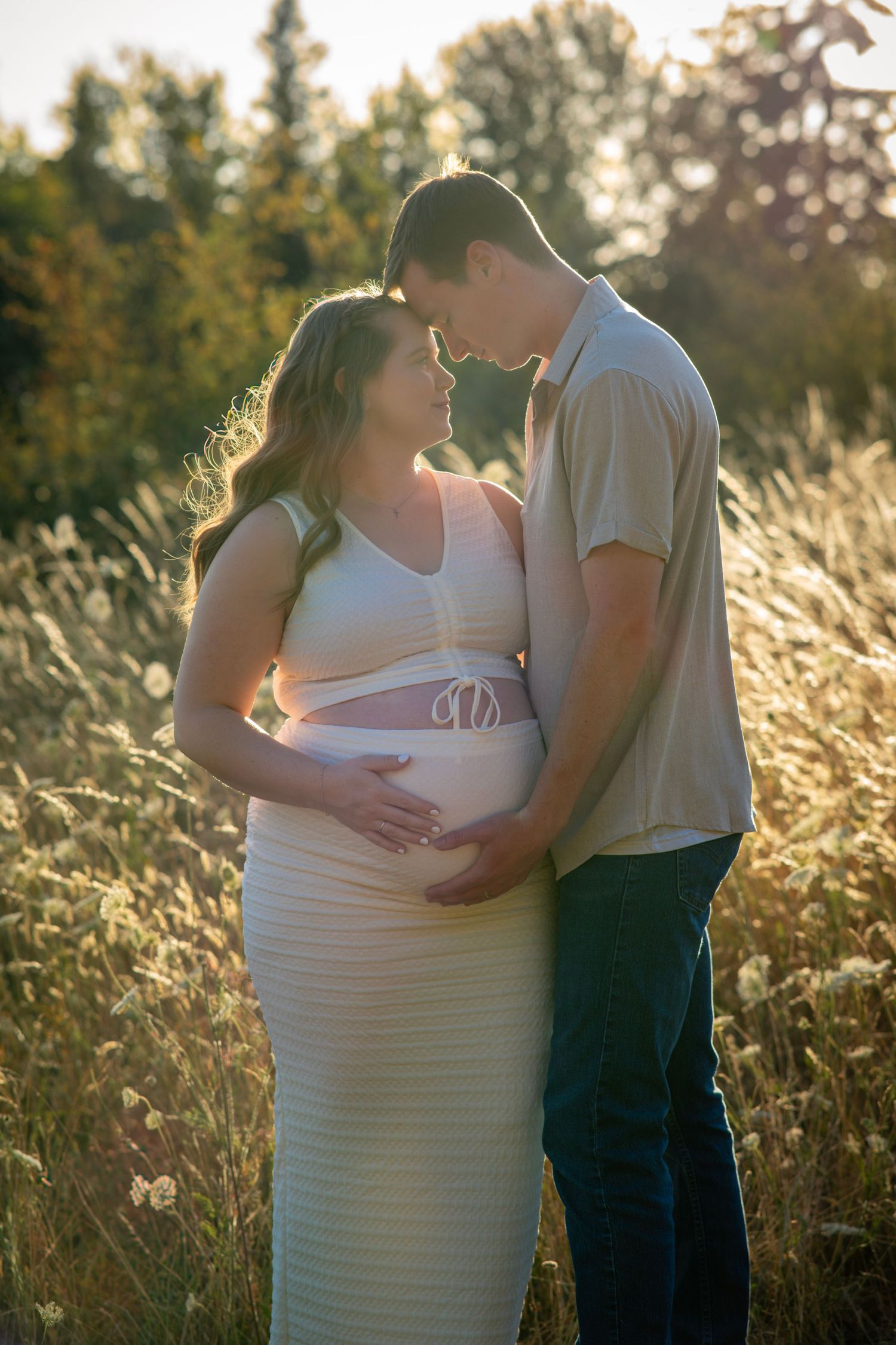 Expectant parents embracing and looking down at the baby bump in warm evening light.