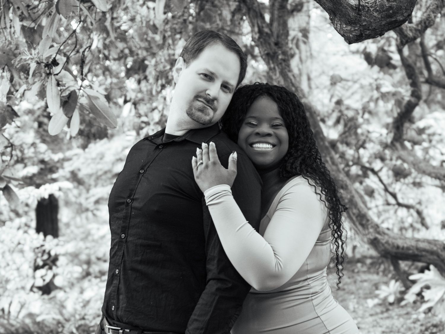 Black and white portrait of a smiling couple embracing beneath leafy trees during an outdoor engagement session.