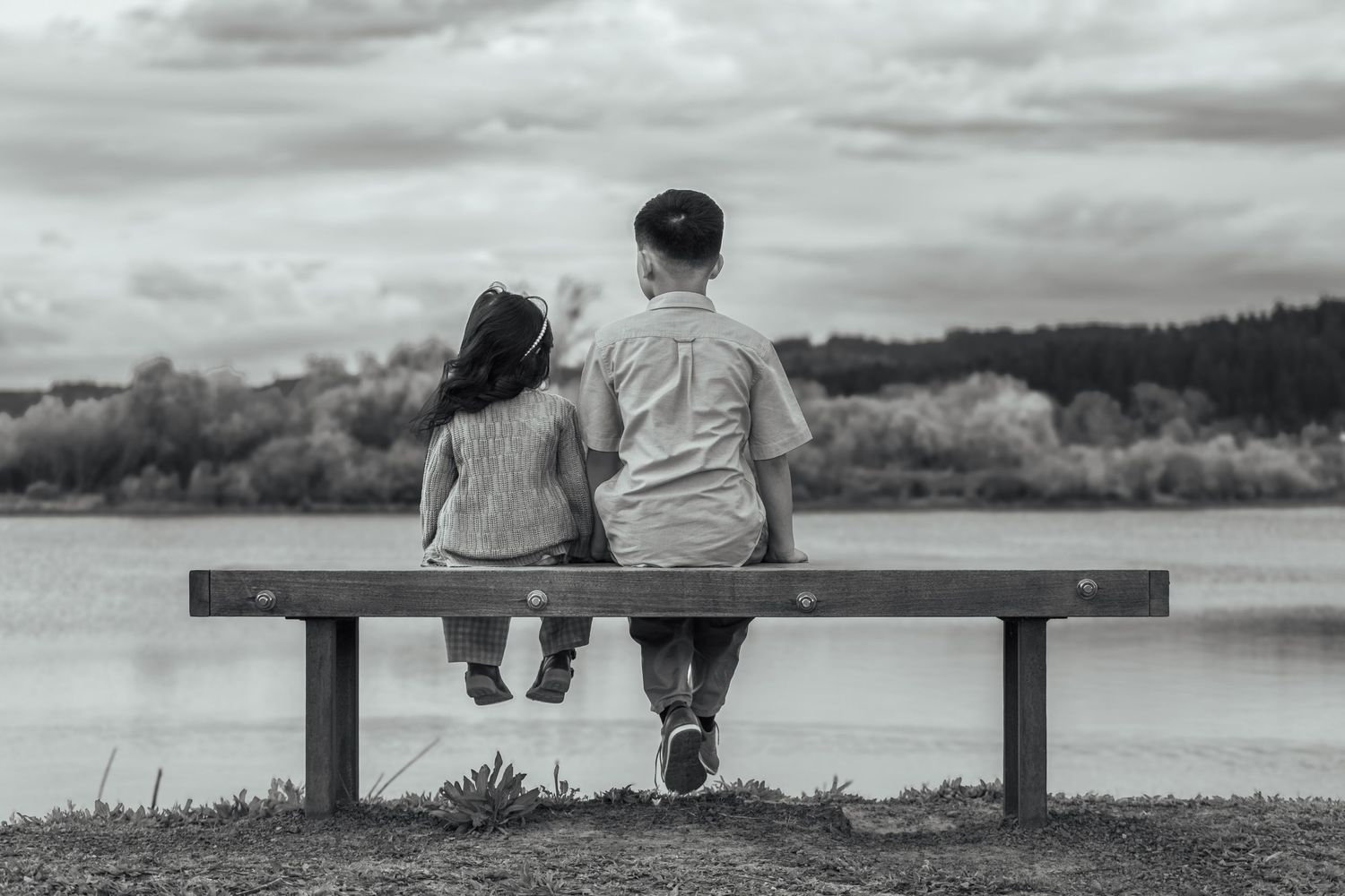Black-and-white candid photo of two siblings sitting together on a bench overlooking the water.