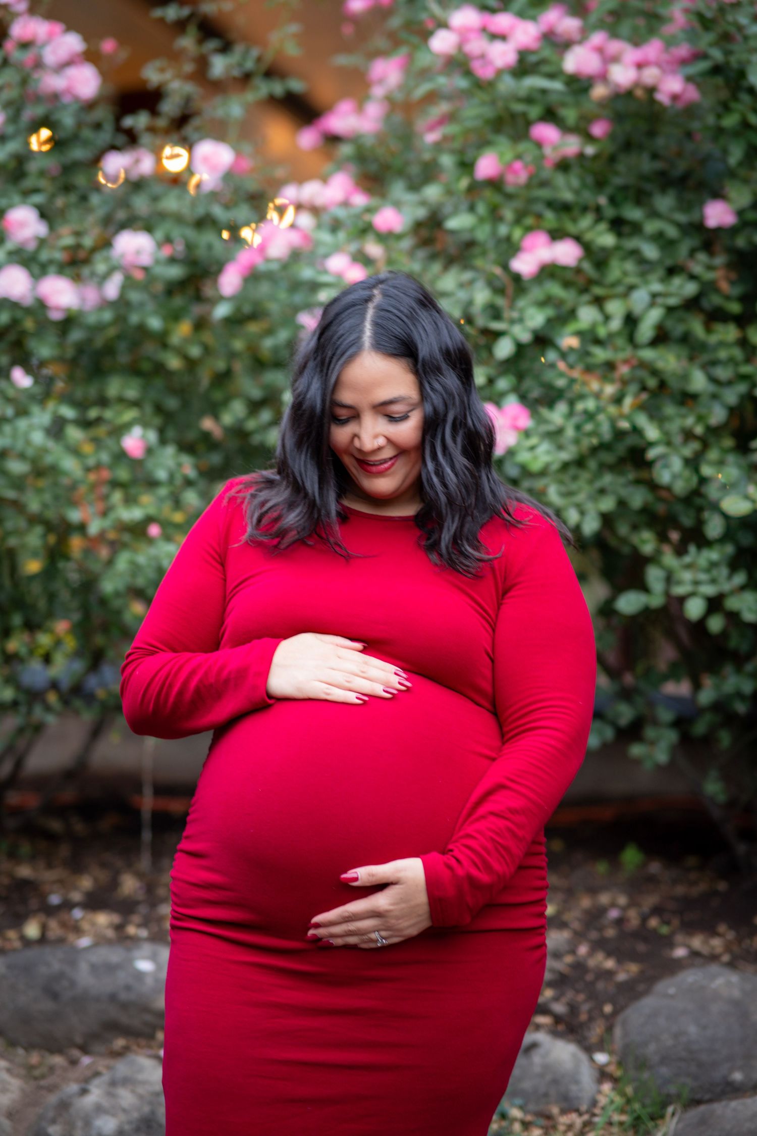 Pregnant woman in a bright red maternity dress looking down at her baby bump in front of blooming pink flowers.
