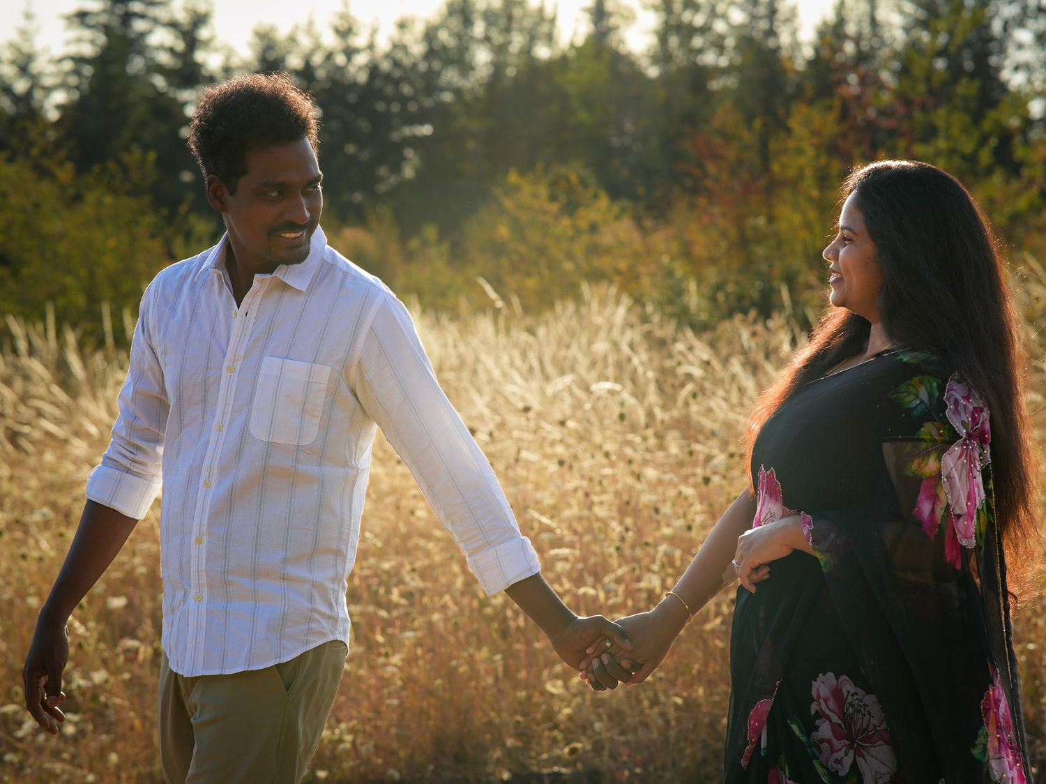 Couple holding hands and looking at each other while walking through a golden field at sunset